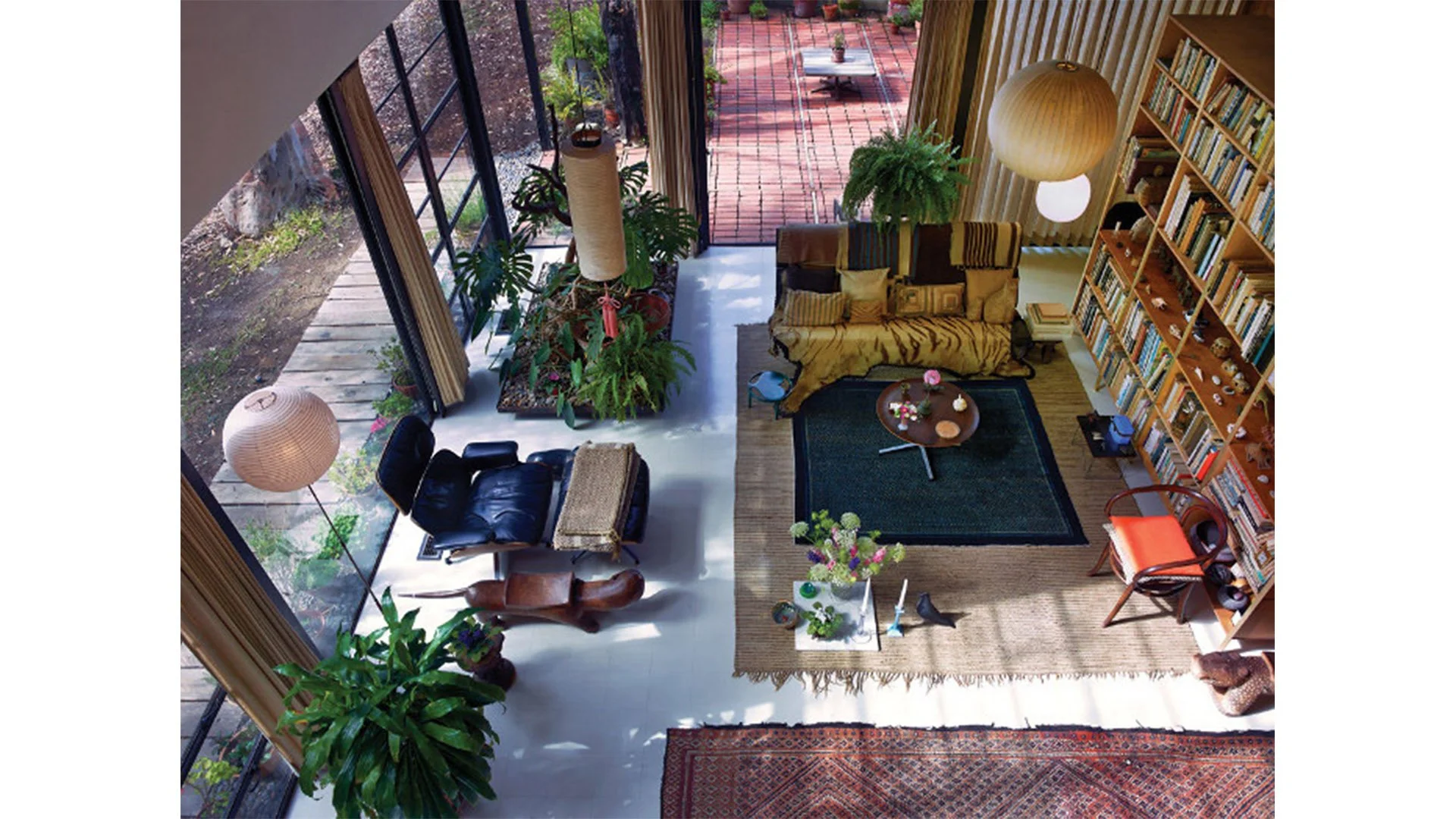 A spacious living room with large floor-to-ceiling windows, greenery outside, a mix of modern and vintage furniture, a large bookshelf filled with books, and a cozy seating area with a patterned sofa, armchairs, and a coffee table decorated with flowers.