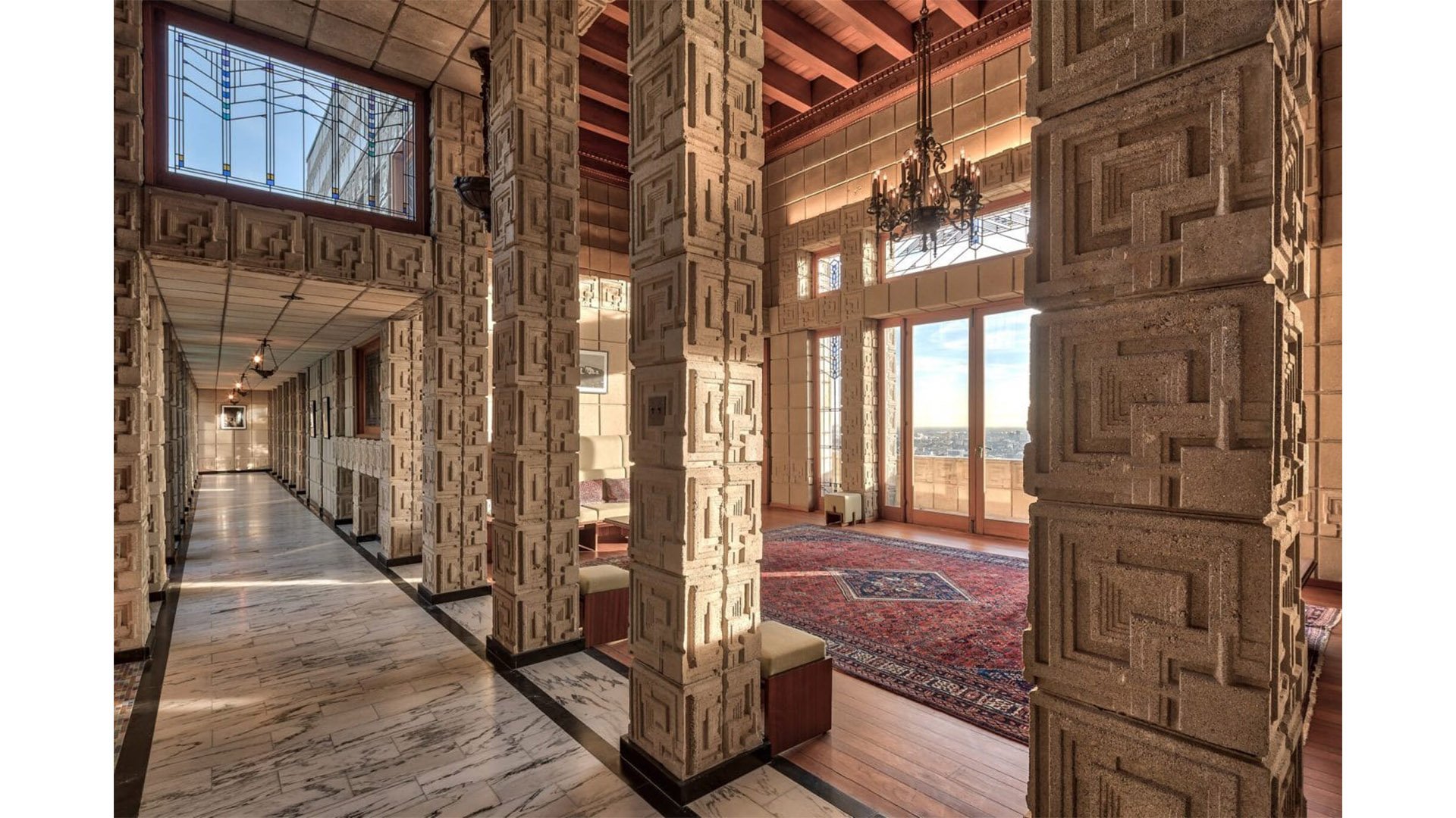 Interior view of a luxurious building featuring intricate stone columns, a marble floor, chandeliers, large windows with a balcony, and a sitting area with a rug.