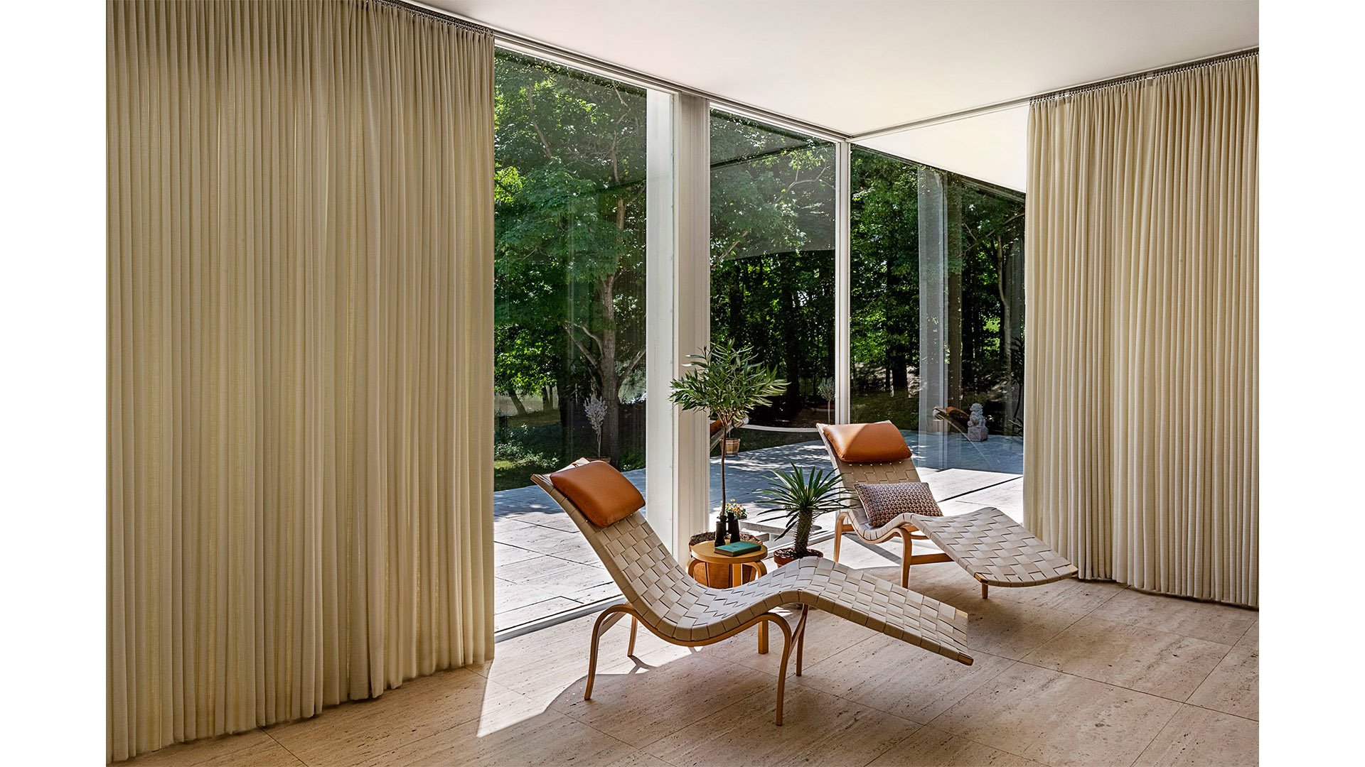 Sunlit interior room with large glass sliding doors, beige floor and curtains, and two mid-century modern lounge chairs with cushions. Potted plants are between the chairs, with an outdoor view of trees and a patio.