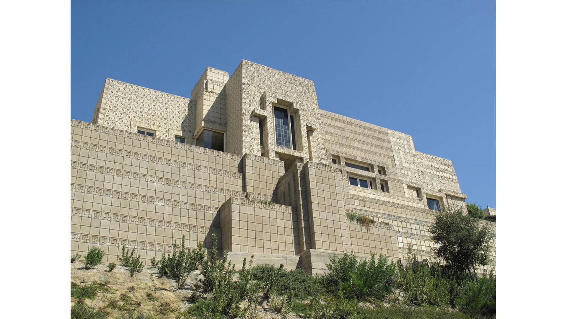 A large, modern beige building with a textured and patterned facade, multiple levels, and rectangular windows under a clear blue sky, with greenery at the base of the building.