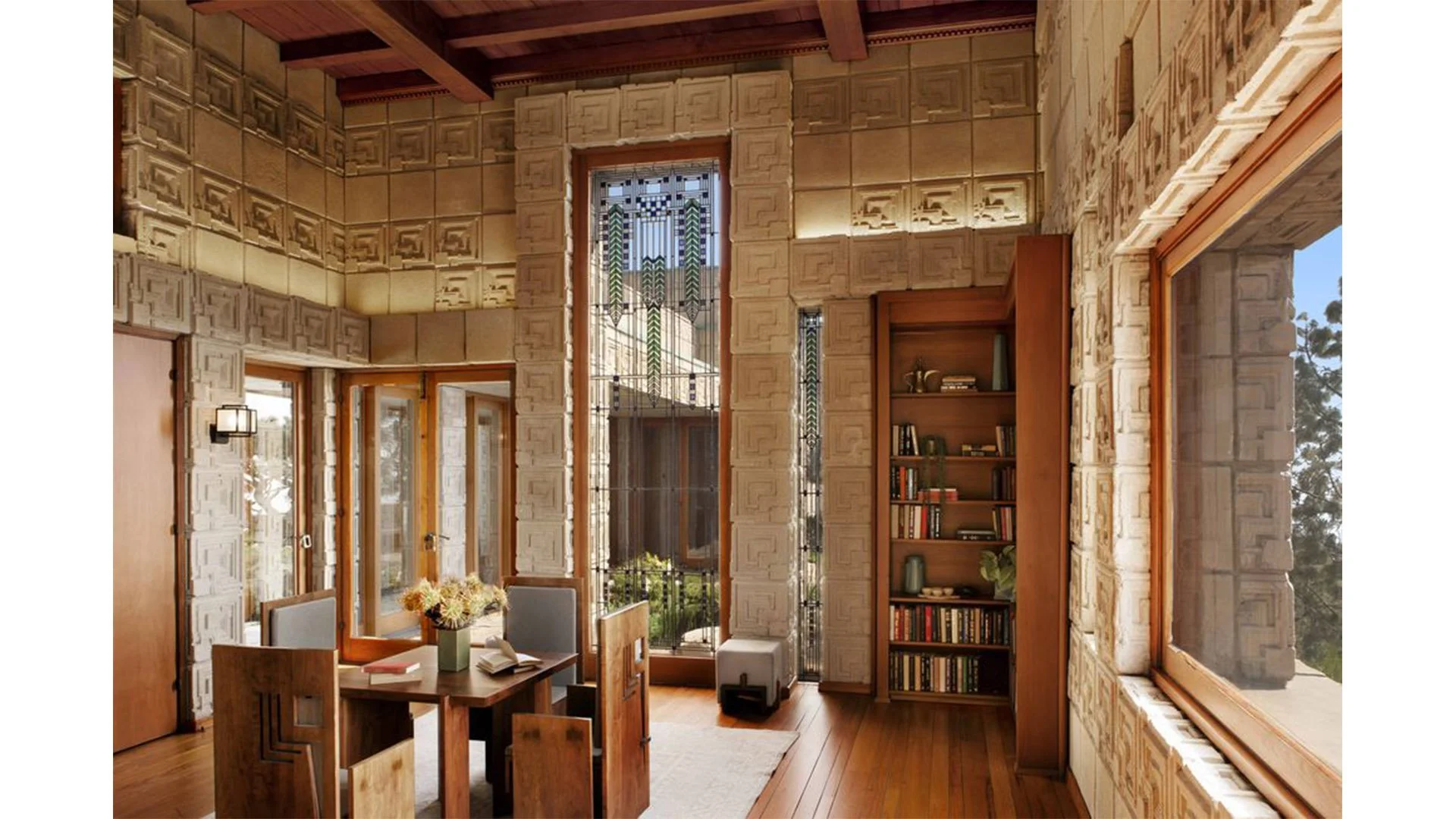 Interior of a room with textured beige stone walls, wooden ceiling beams, a window, glass door, bookshelf, table with chairs, and a vase with flowers.