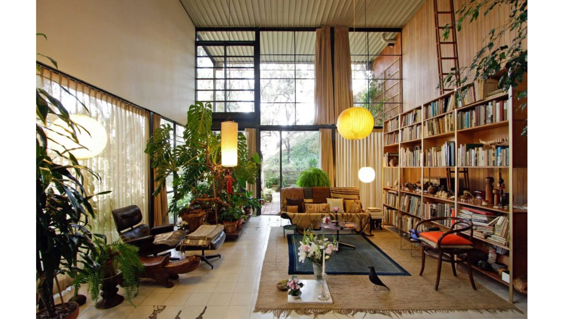 Living room with large windows, wooden walls, bookshelves, sofas, plants, and hanging lamps.