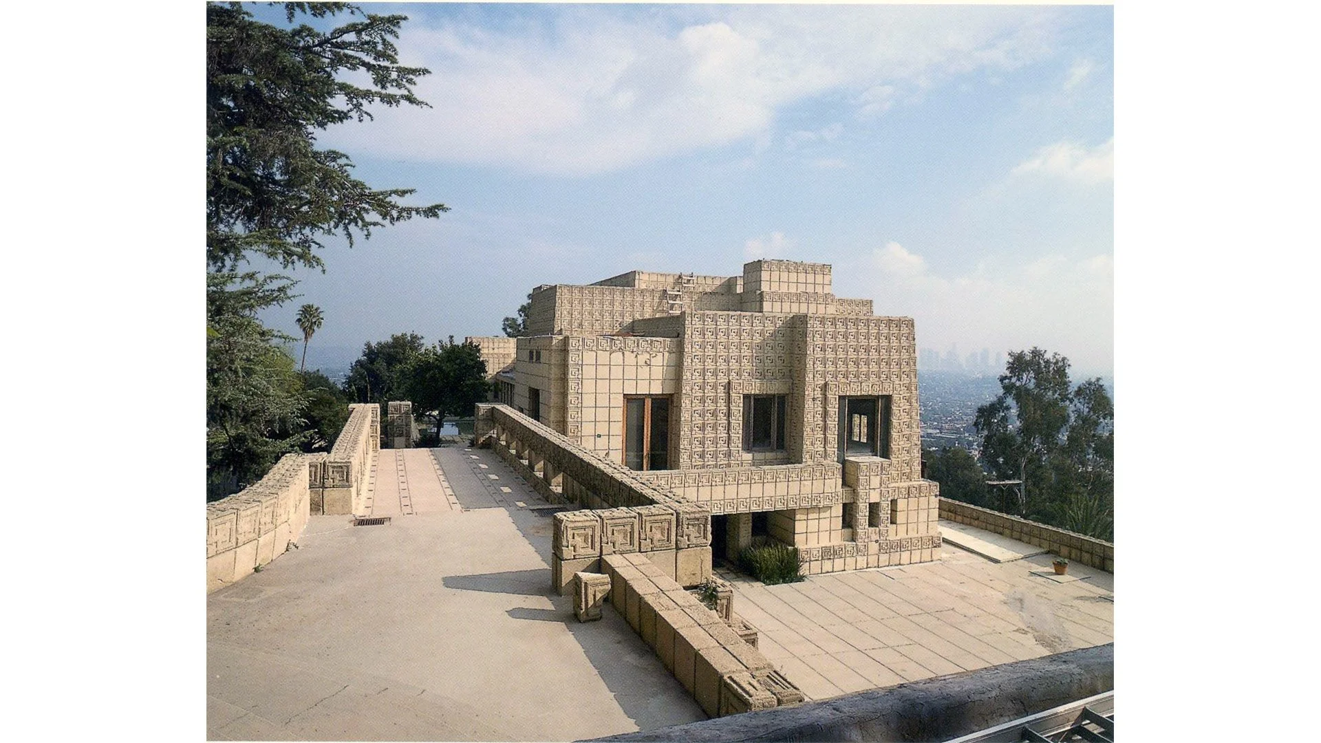 A beige building with an intricate geometric pattern, set on a hilltop with a city skyline in the background. There are trees and a clear blue sky.