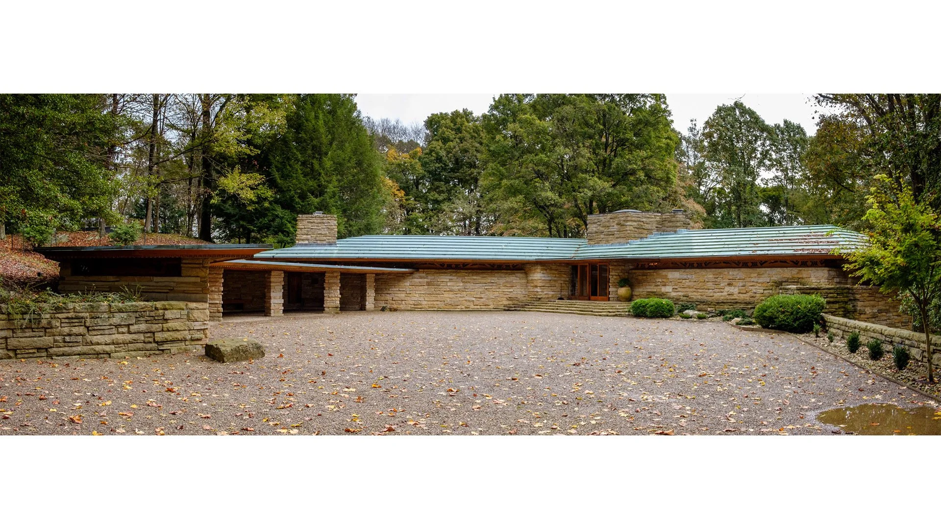 A modern house with stone walls and a unique sloped roof surrounded by trees and a gravel driveway.