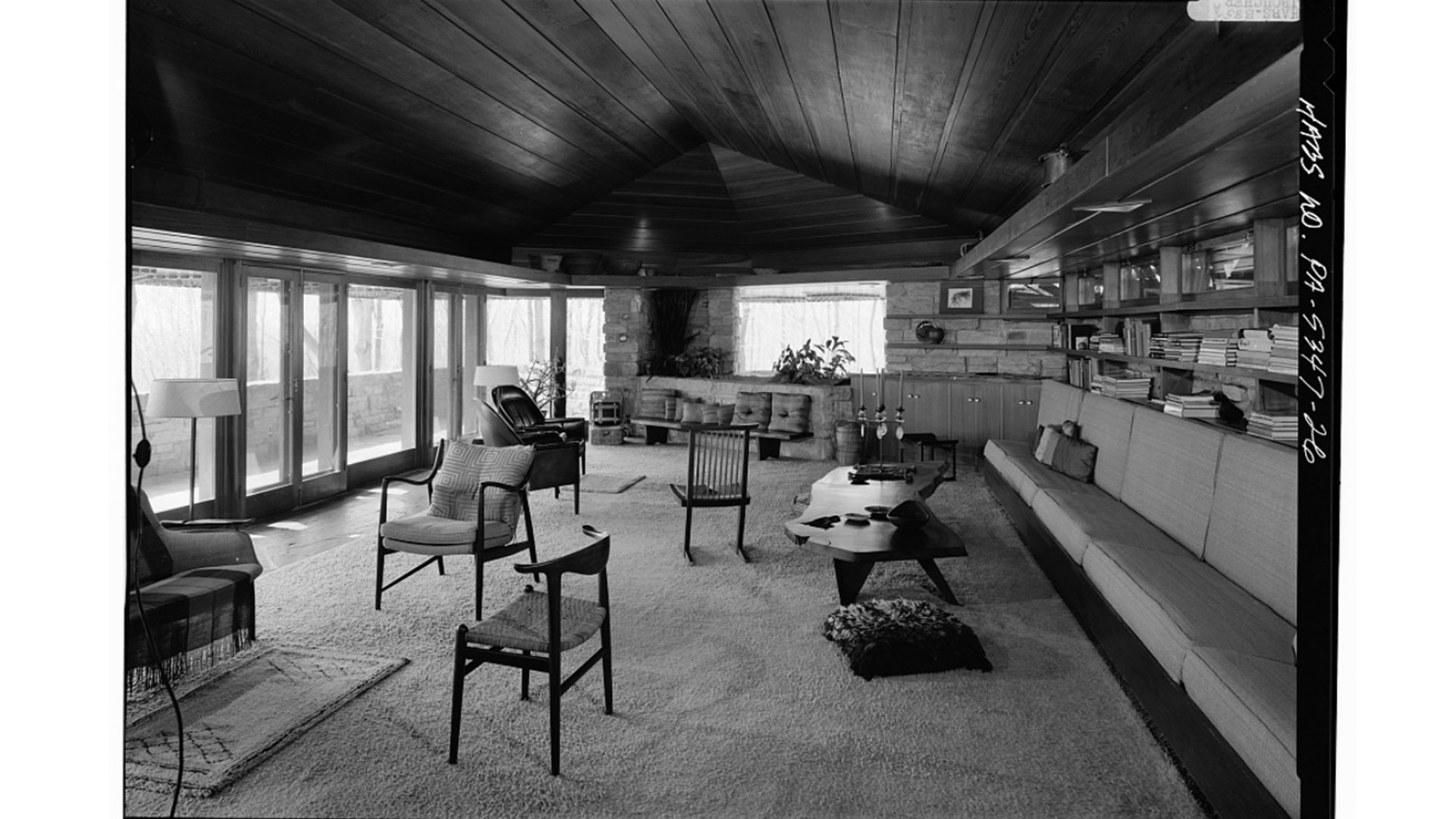 A living room with large windows, wooden ceiling, and a built-in sofa against the wall lined with books. Various chairs, a coffee table, and a floor lamp are also present.