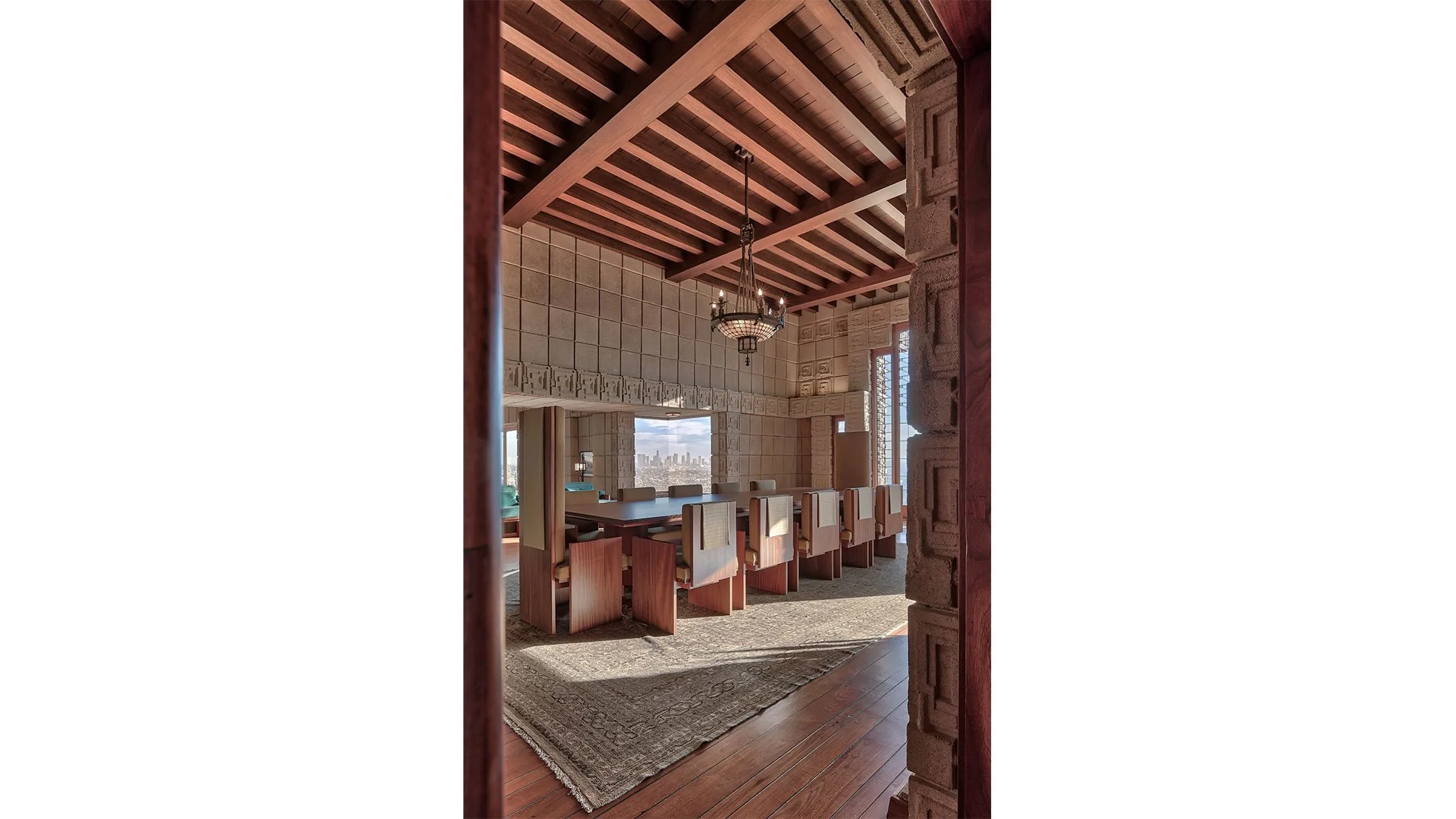 Interior of a cozy dining room with a large dark wood table and cream upholstered chairs; a chandelier hangs from a wooden ceiling with exposed beams; large windows offer a view of a city skyline in the distance.
