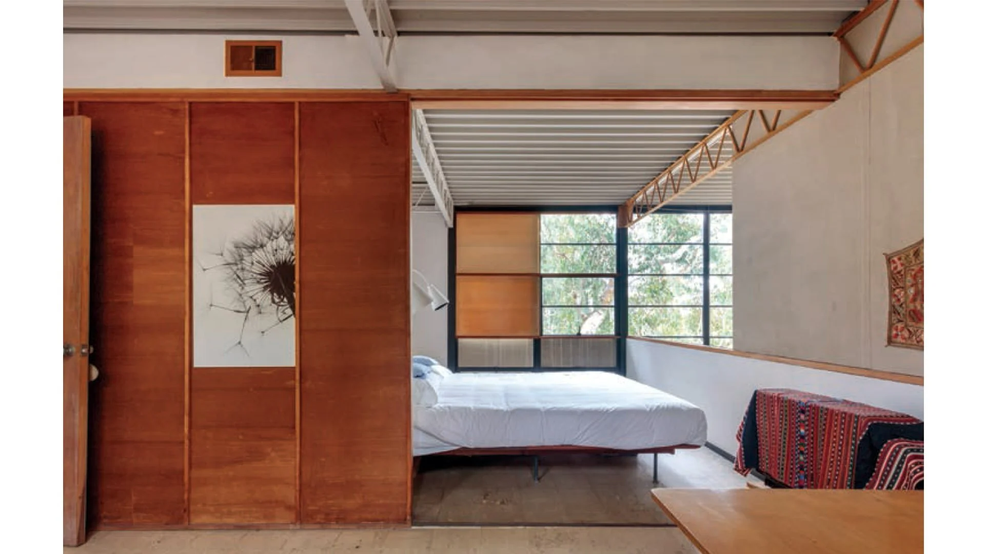 A bedroom with a bed, a large window, wooden closet doors, and a colorful patterned chair next to a small wooden table.