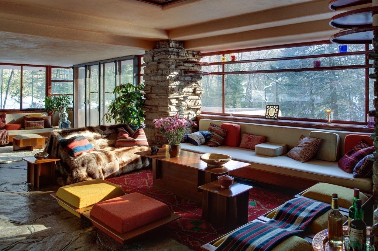 A cozy living room with large windows showing a snowy outdoor scene, featuring a stone column, a mix of colorful seating including a fur-covered couch, a long built-in bench with many pillows, and various small tables with decorative items and bottles.