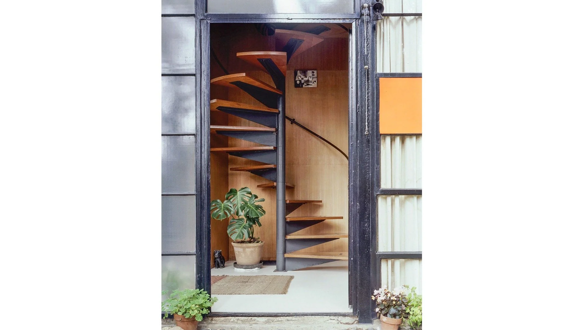 View through a glass door showing a spiral staircase with black and wooden steps, a potted Monstera plant, a small black cat figurine, and a framed black-and-white abstract art piece on the wall.