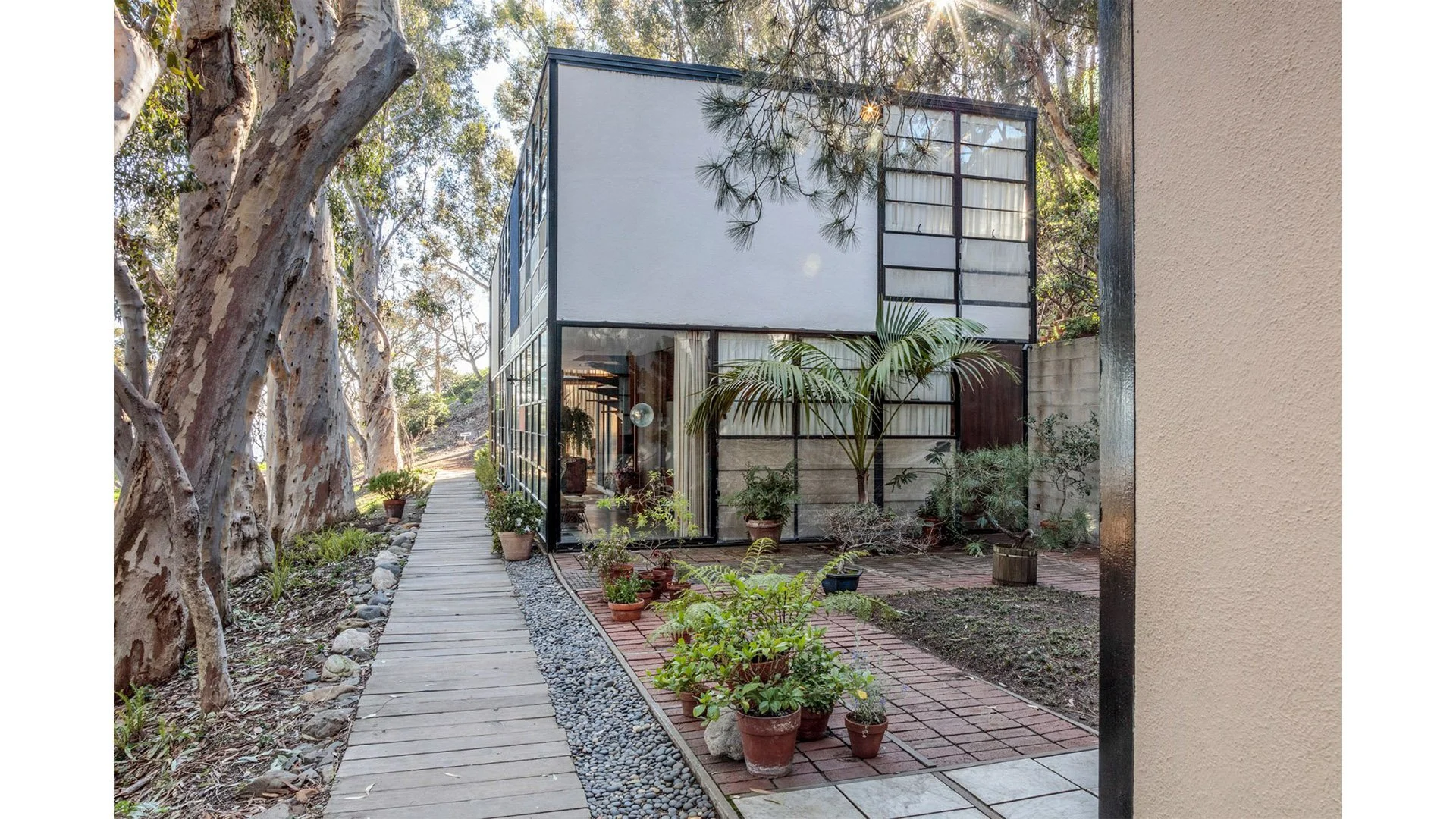 A modern house with large glass windows, surrounded by potted plants and trees, with a wooden walkway and outdoor garden area.
