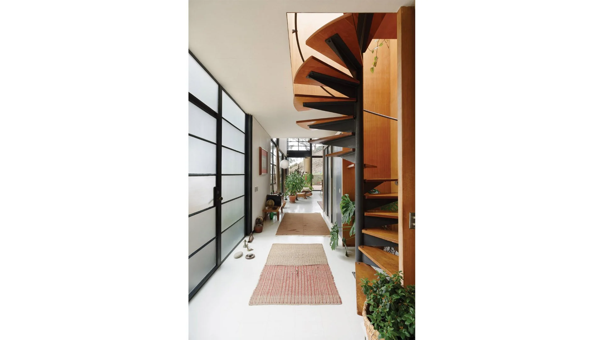 Interior hallway with black-frosted glass sliding doors on the left, a spiral staircase with wooden steps and black support on the right, several rugs on white floor, and plants at the end of the hallway leading to an outdoor space.