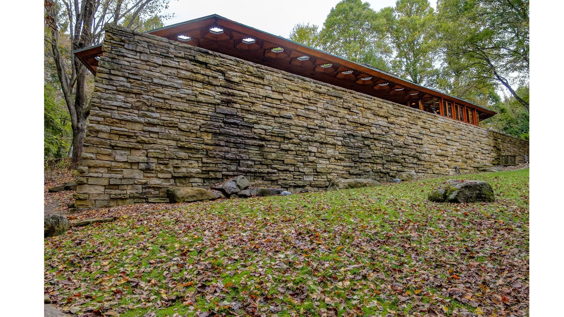 A large stone retaining wall on a sloped lawn with fallen autumn leaves. The wall is built with rectangular stones and has a wooden deck or porch with a roof on top, set amidst trees.