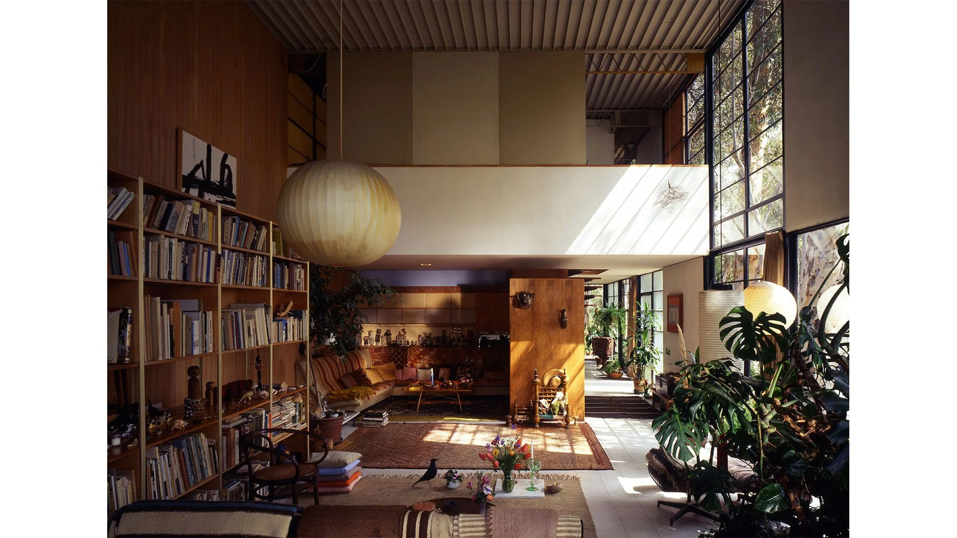 Interior of a living room with large windows, a bookshelf filled with books, a hanging paper lantern, potted plants, seating area, and sunlight streaming through the windows.