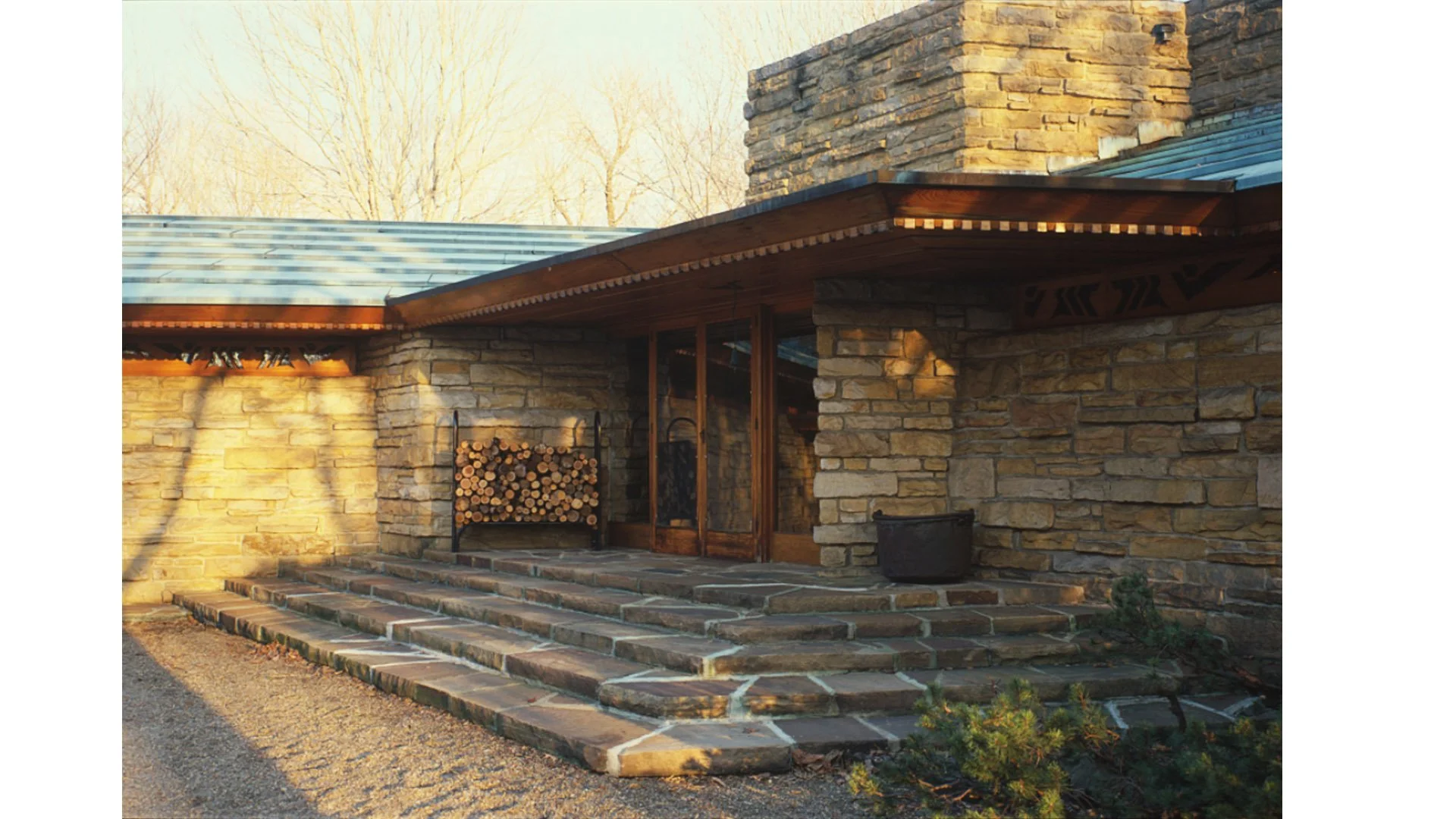 Front view of a stone house entrance with a small staircase, a wood door with glass panels, and a firewood rack next to the door, with sunlight casting shadows on the stone walls.