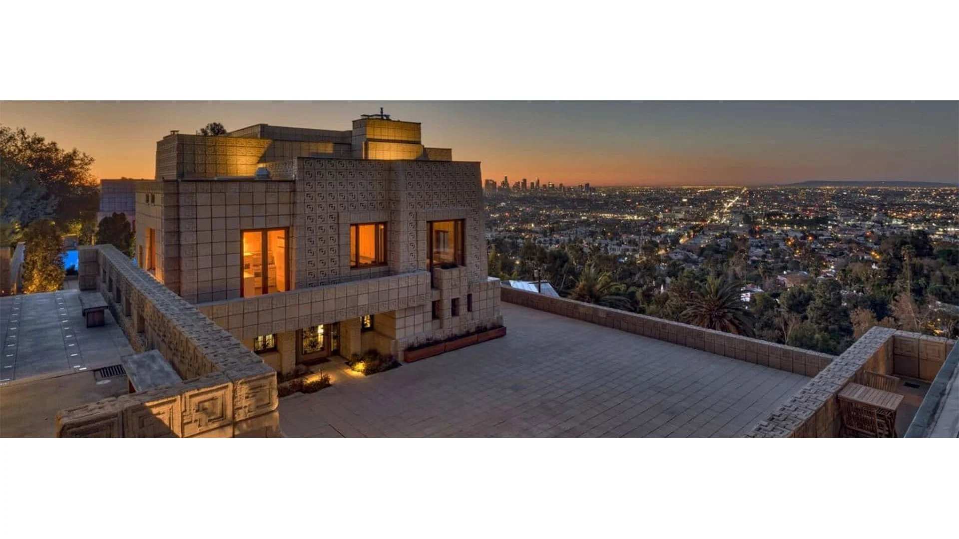 View of a rooftop terrace overlooking a city skyline at sunset with illuminated buildings and trees.