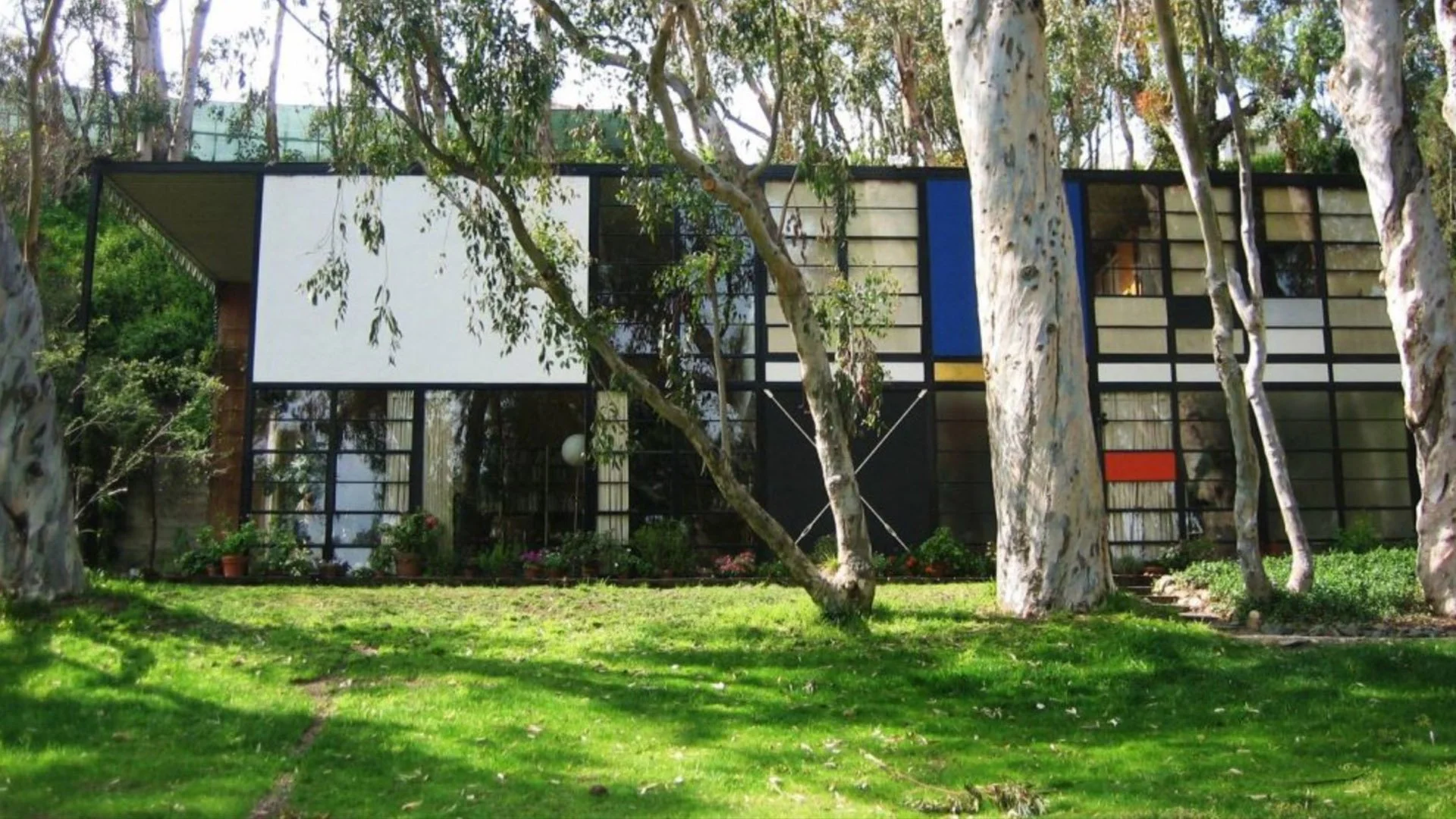Modern house with large black-framed windows and multicolored geometric panels, surrounded by trees and a green lawn.