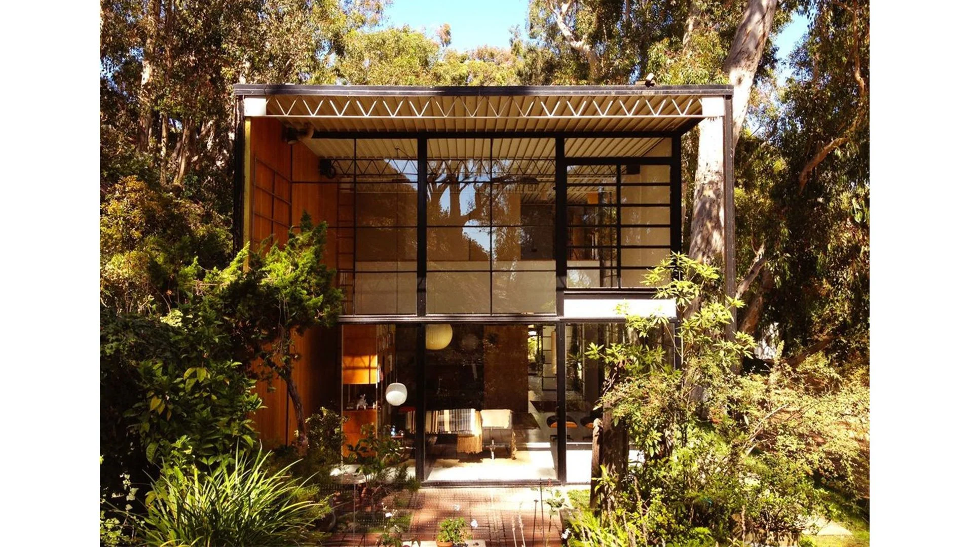 A modern two-story house surrounded by lush greenery, with floor-to-ceiling black-framed glass windows and a brick pathway leading to the entrance.
