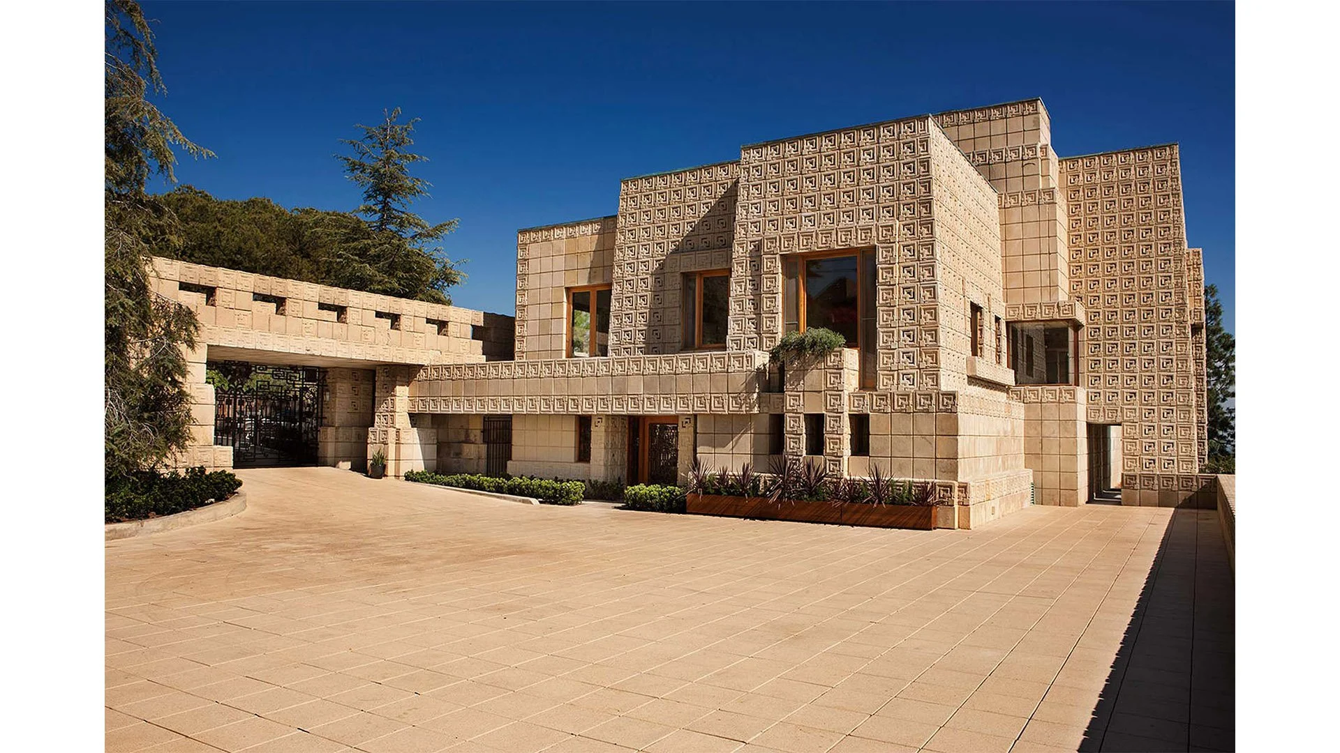 Modern beige stone building with geometric patterned walls, large windows, and a gated entrance, surrounded by landscaped garden and trees, under a clear blue sky.