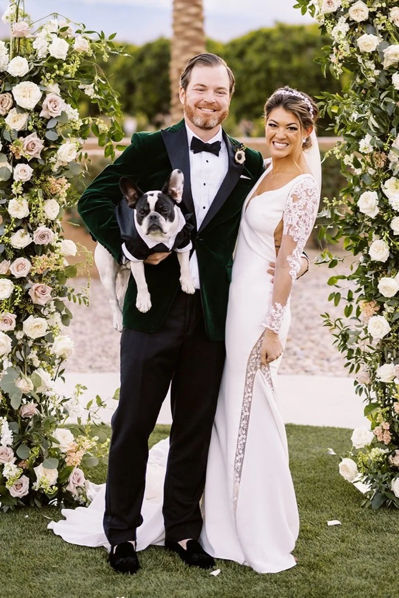 A bride and groom standing together at their wedding ceremony outdoors, framed with floral arrangements, with a French bulldog in formal attire, greenery, and a clear sky in the background.