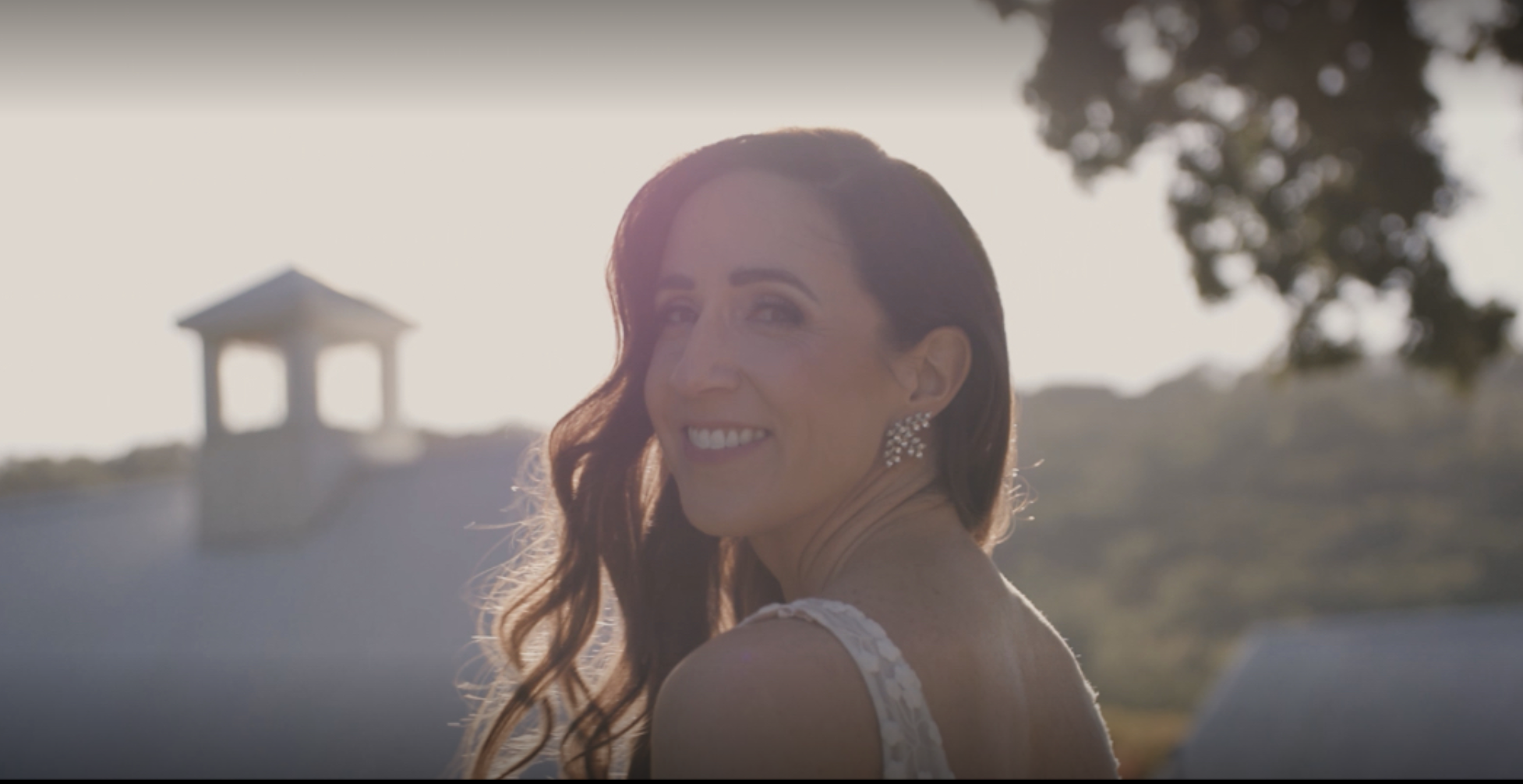 A woman with long auburn hair smiling outdoors during sunset, wearing a white dress and sparkling earrings.