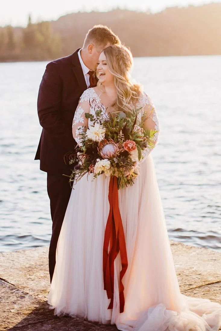 A happy couple in wedding attire sharing a joyful moment by a lake at sunset. The bride is holding a large bouquet of flowers.