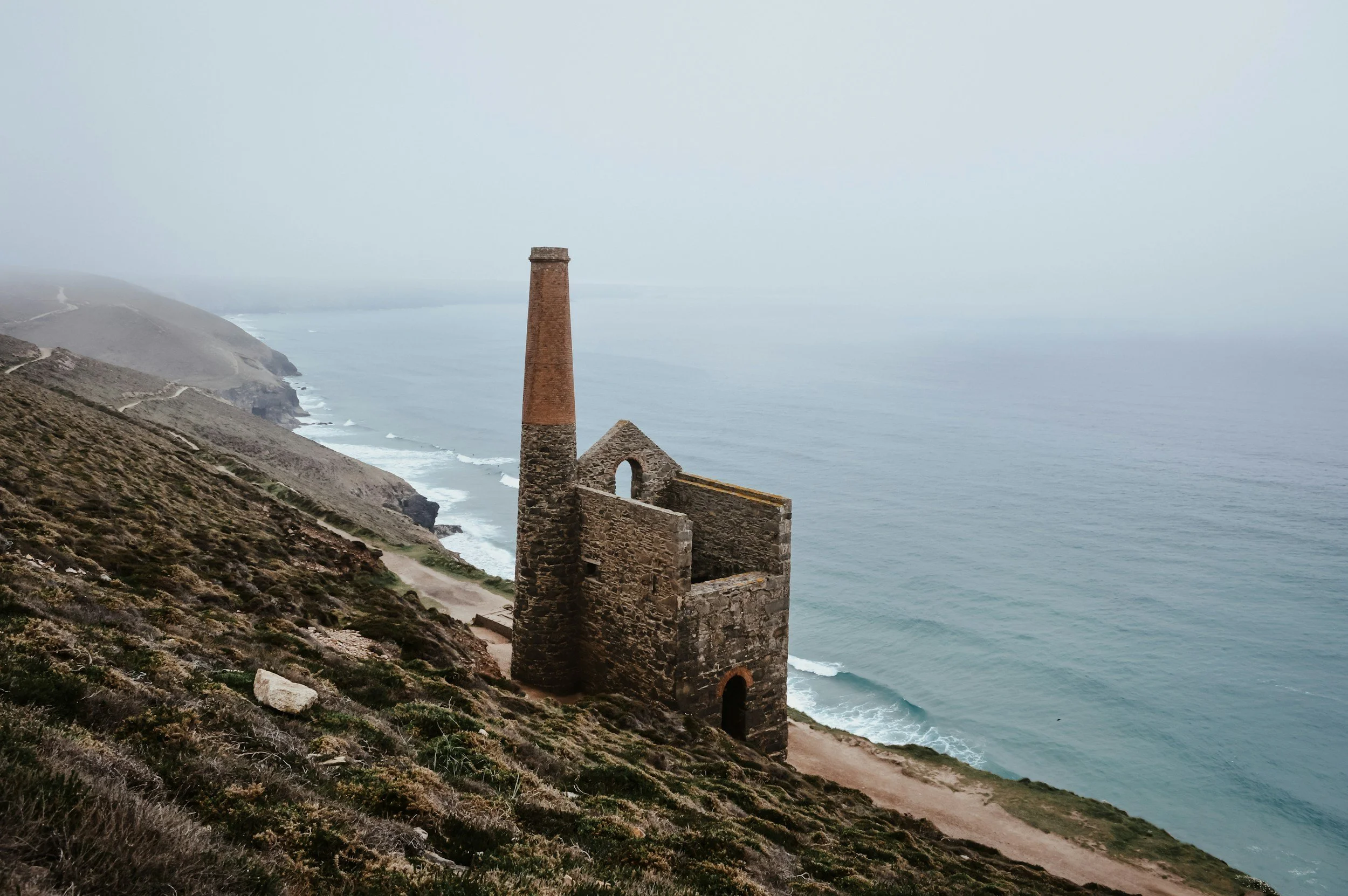 An abandoned stone building with a tall chimney on a foggy coastal hillside overlooking the ocean.