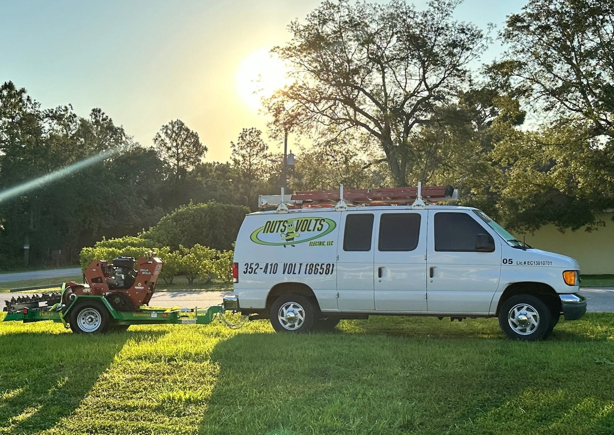 A utility van parked on grass with tools on roof rack and a trailer hitched to it, carrying equipment for tree trimming or similar work, with trees and a setting sun in the background.