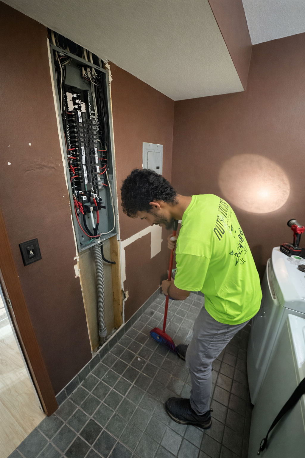 A man in a bright yellow shirt sweeping the floor of a laundry room next to an open electrical panel on the wall.