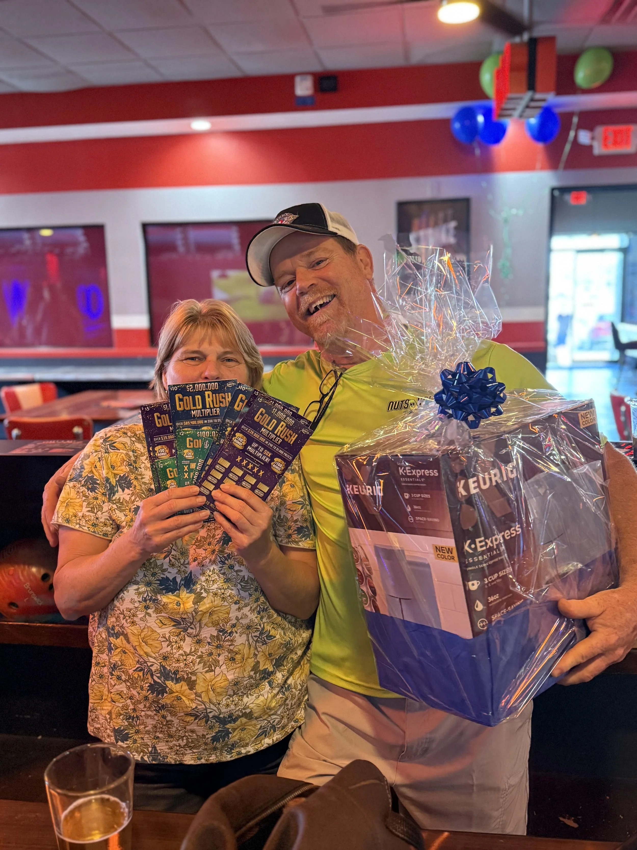 A woman and a man celebrating at a restaurant, holding lottery tickets, with the man holding a large gift basket wrapped in plastic. The woman is smiling and holding multiple lottery tickets.
