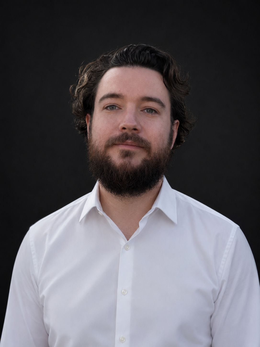 Portrait of a young man with dark curly hair and a beard, wearing a white dress shirt, against a black background.