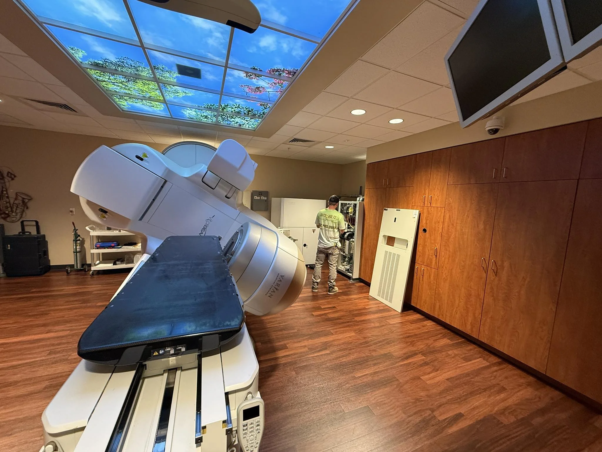 A medical imaging room with a large MRI machine, a technician working on equipment, a wooden cabinet, and a ceiling with illuminated nature-themed panels.