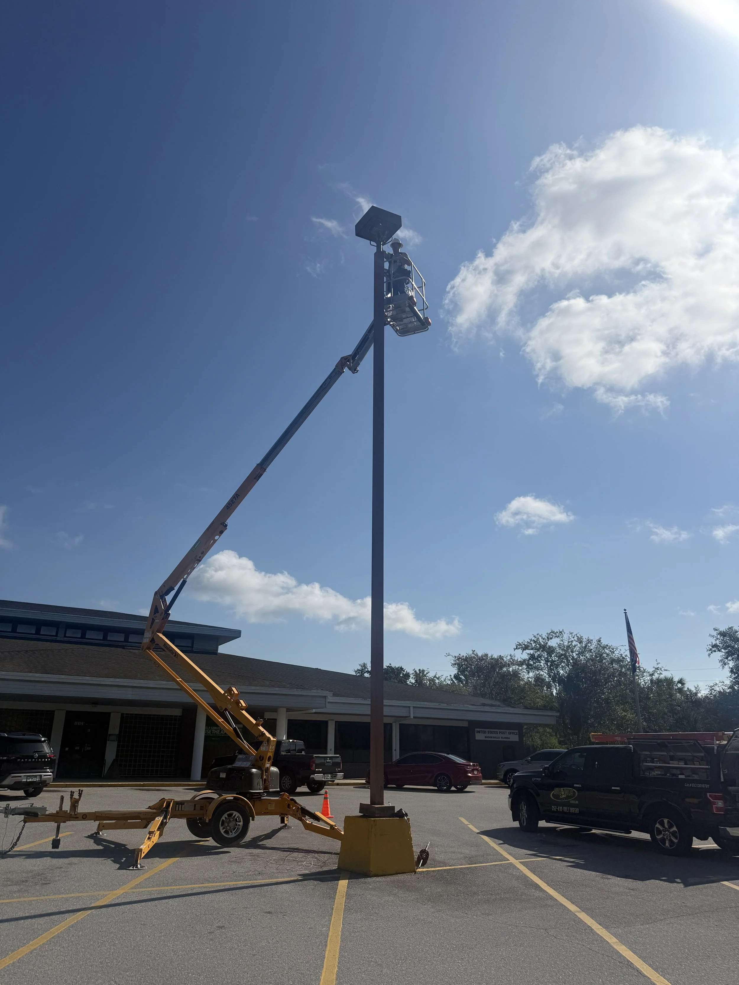 A person in a bucket lift working on a tall pole in a parking lot with several cars and a building with a United States Post Office sign in the background.
