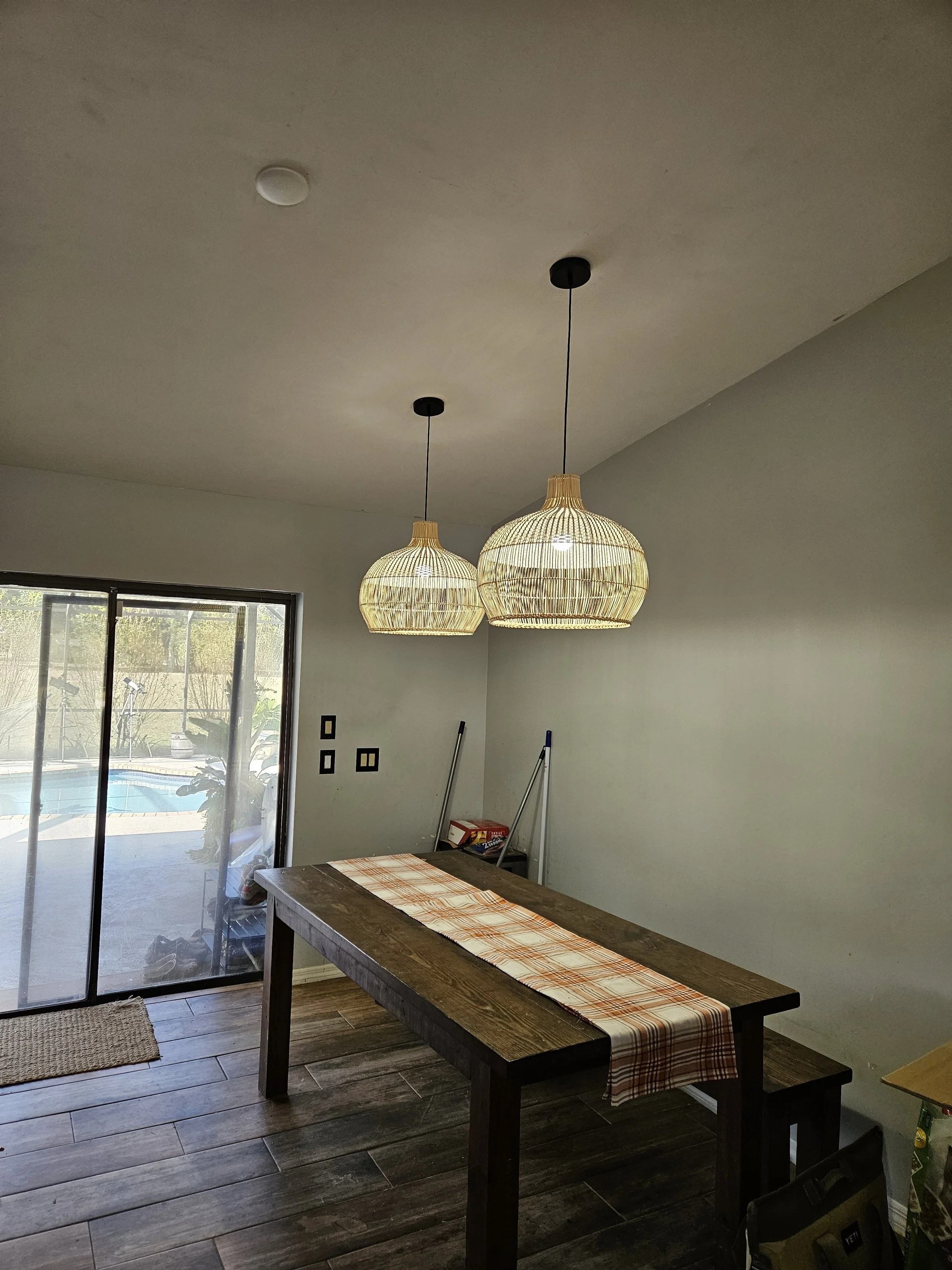 A dining room with two wicker pendant lights, a wooden table with a plaid table runner, sliding glass door leading to outside, and some cleaning tools in the corner.