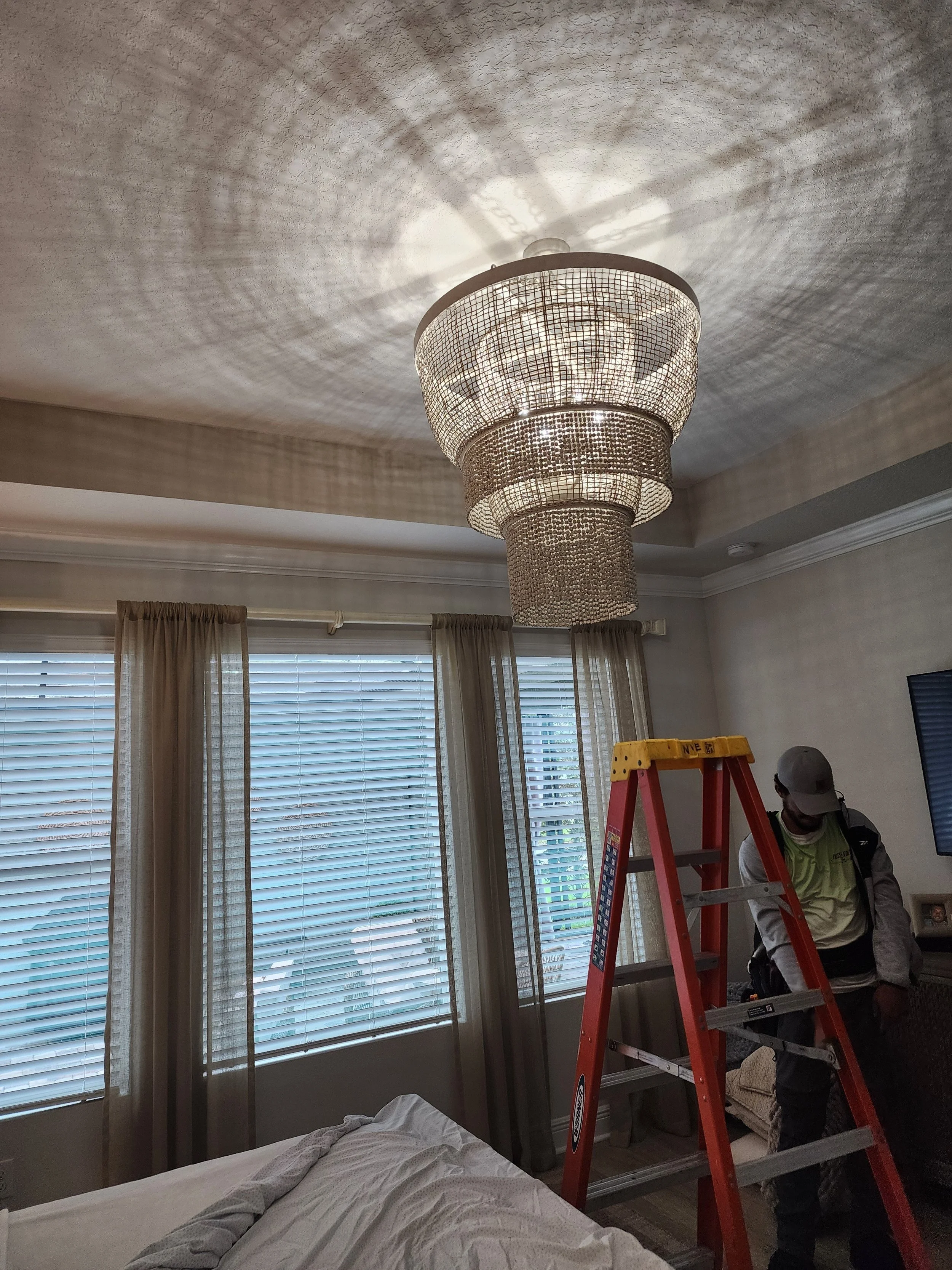 Person standing on a ladder in a living room near a large chandelier, with window curtains and a television on the wall.