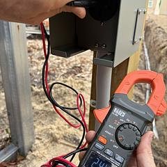 Person using digital multimeter to measure electrical connections outdoors, with wires attached to a metal pole.
