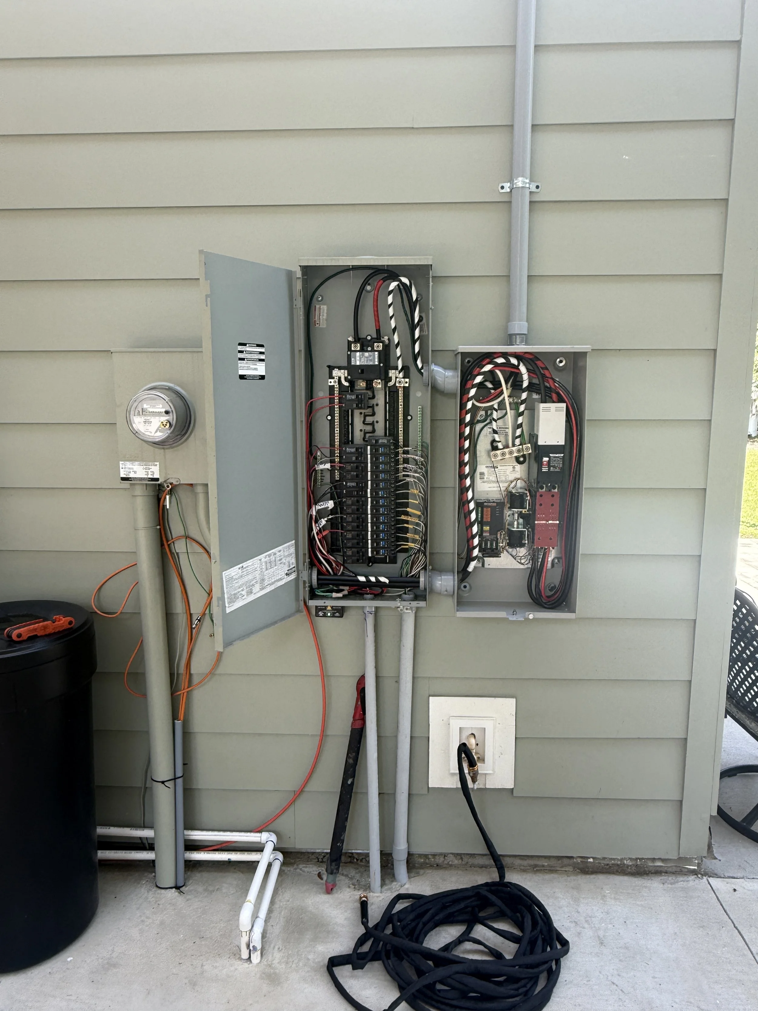 An outdoor electrical panel with open doors revealing wiring and circuit breakers, set against a beige house siding wall, with conduit pipes, a black water filter, and coiled black hose on the ground.