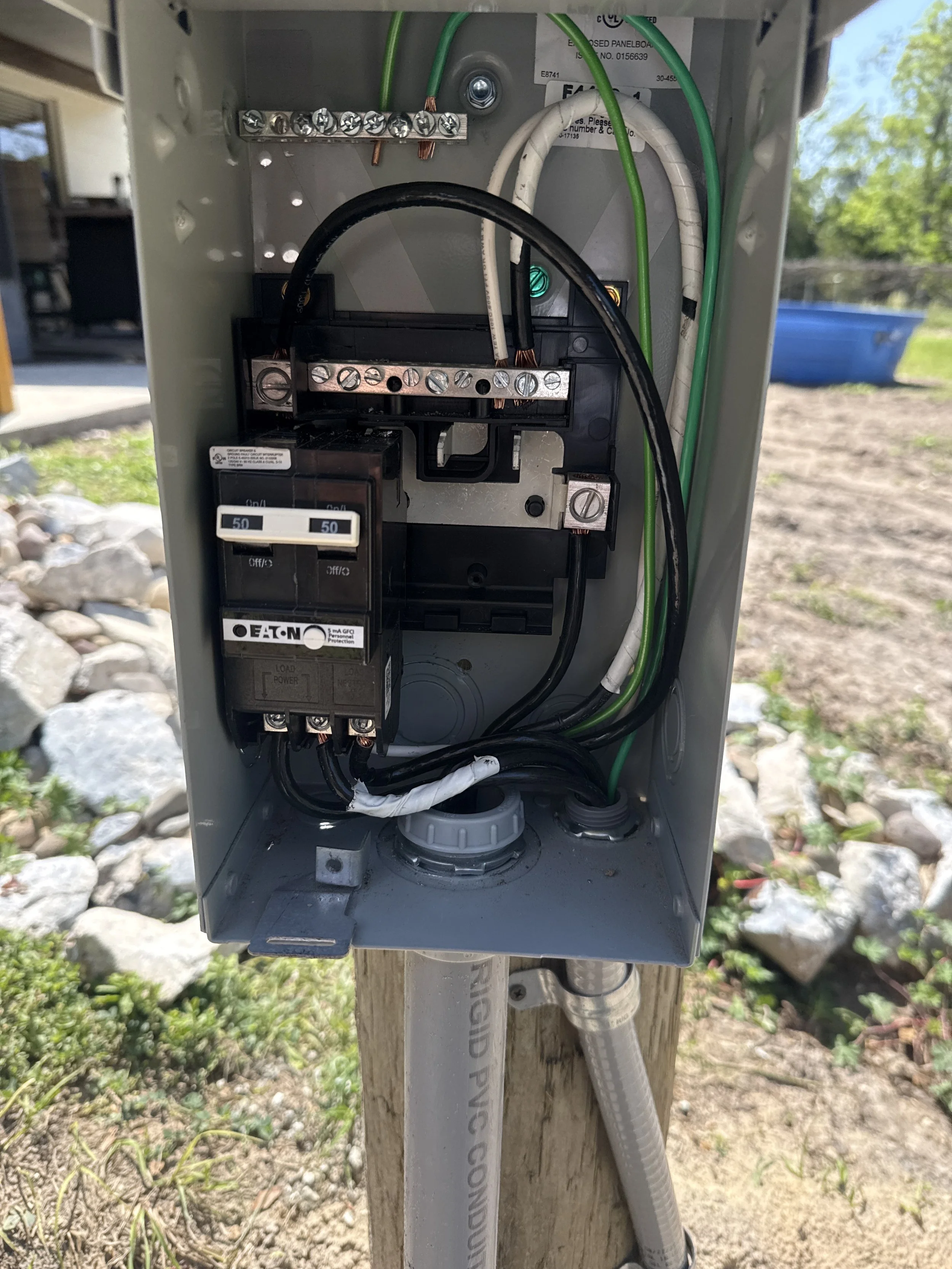 Open electrical box on a wooden pole with colored wires and a circuit breaker, set outdoors on rocky ground with grass and trees in the background.