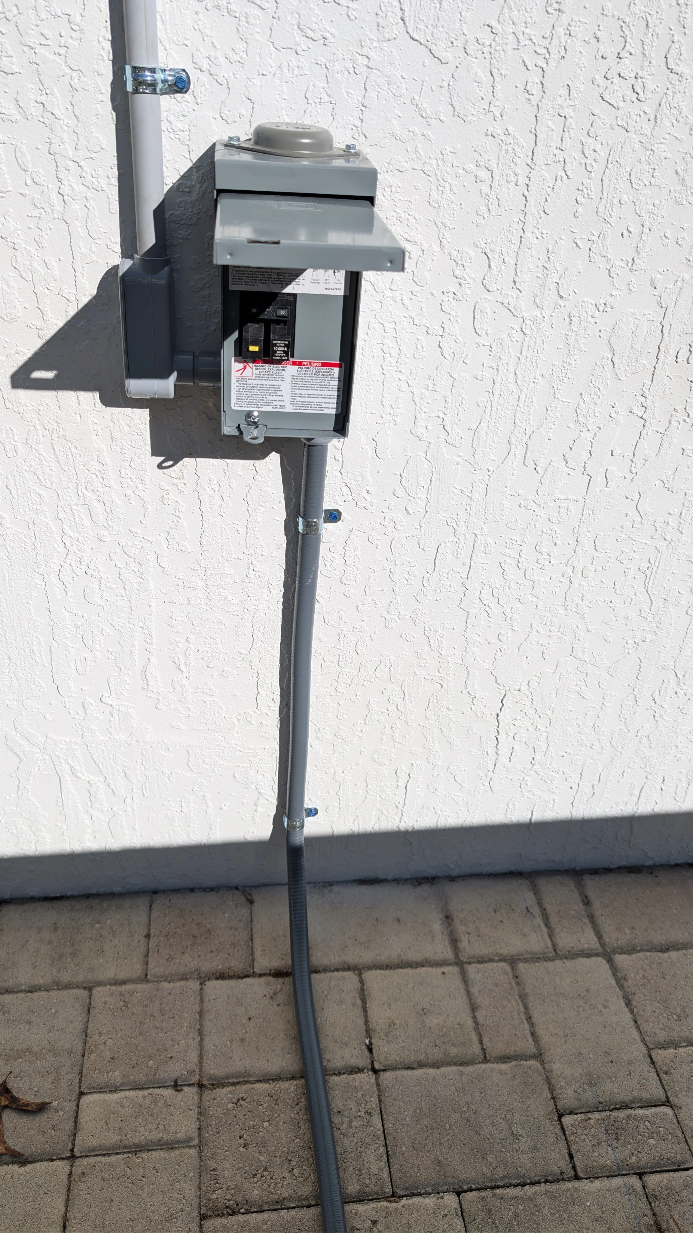 An electrical meter box, mounted on a wall exterior, with conduit piping and a digital meter, casting a shadow on the textured wall.