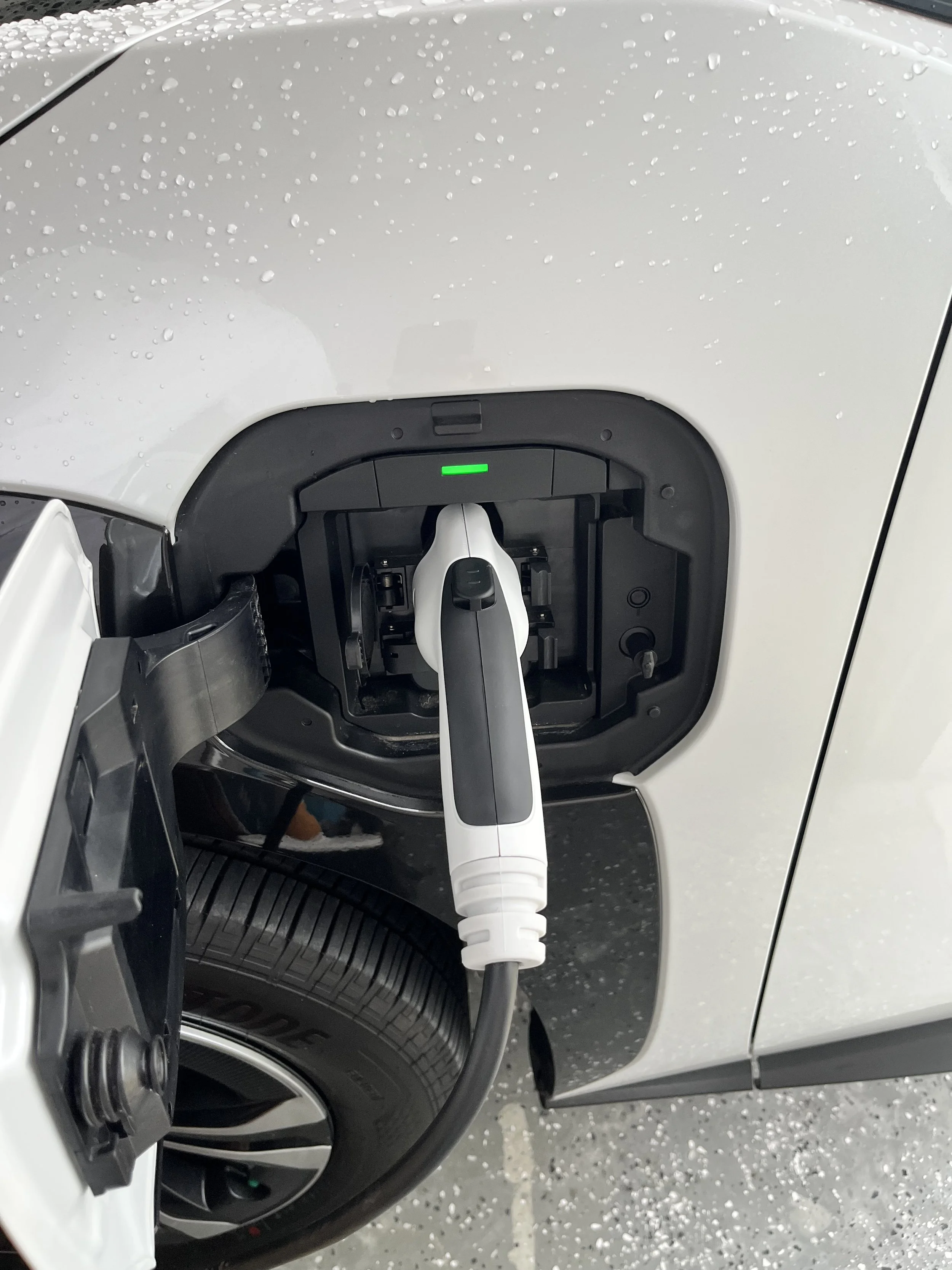 Electric vehicle charging port with an attached charging cable, located on the side of a white car with raindrops on the surface.