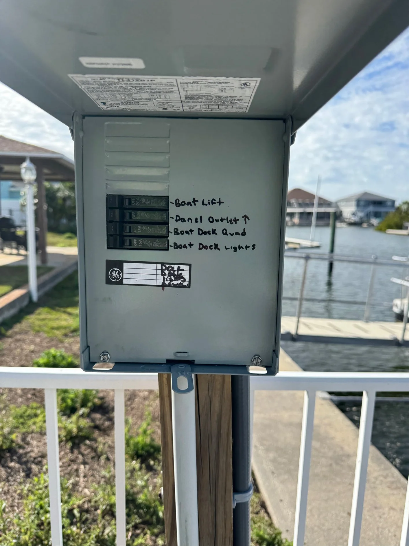 An electrical panel with handwritten notes indicating the circuit functions, including boat lift, panel outlet, boat deck ahead, and boat deck lights, located near a harbor with houses and boats in the background.