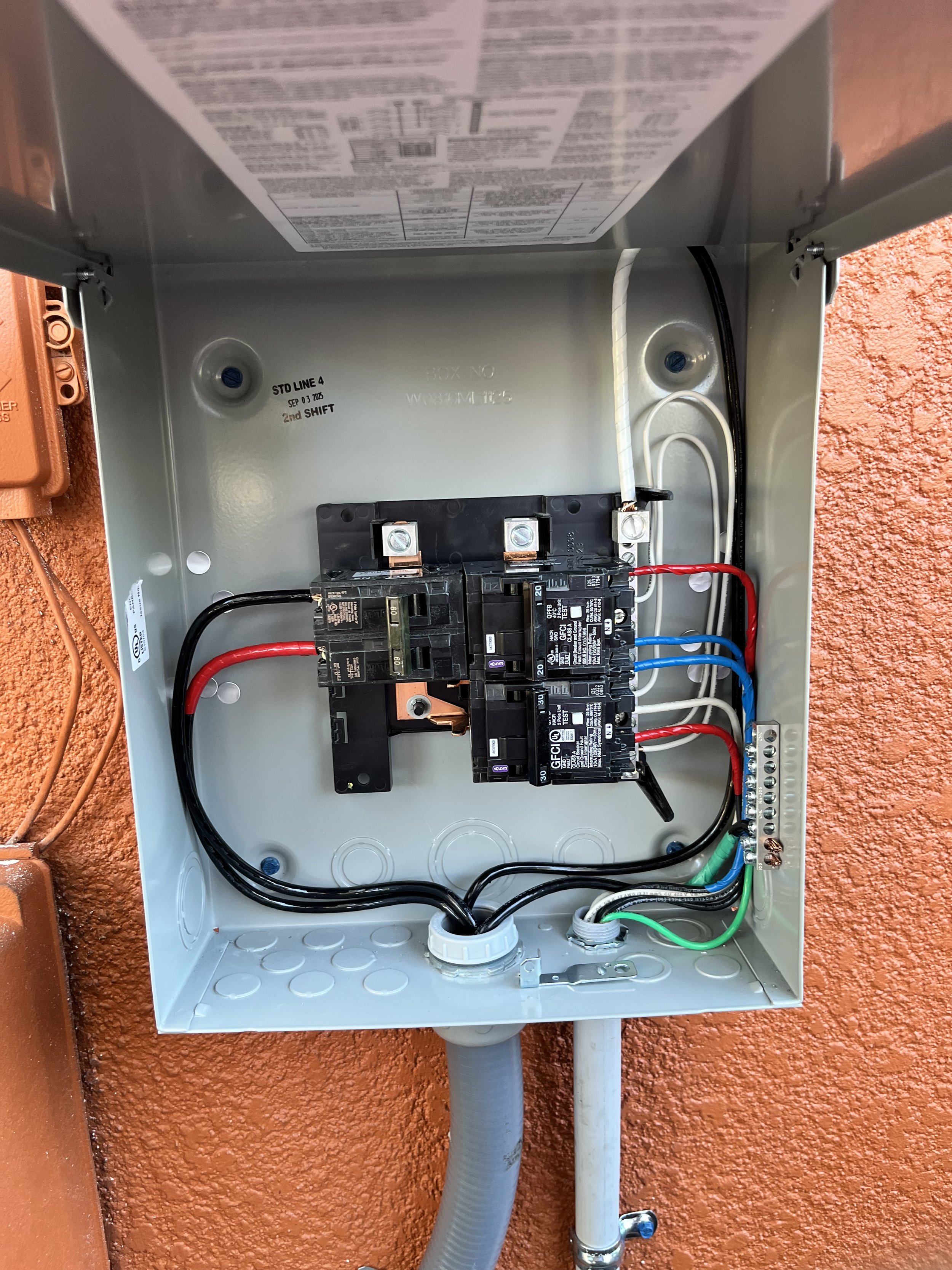Open electrical meter box with circuit breakers and wiring inside, mounted on an orange wall.