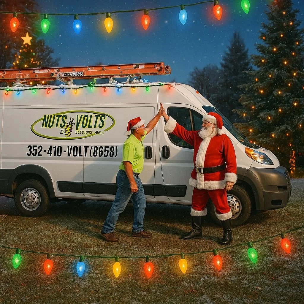 A man dressed as Santa Claus and another man in a neon green shirt and jeans giving each other a high-five in front of a company van labeled 'Nuts & Volt.' They are standing outdoors at night decorated with Christmas lights and a large illuminated Christmas tree in the background.
