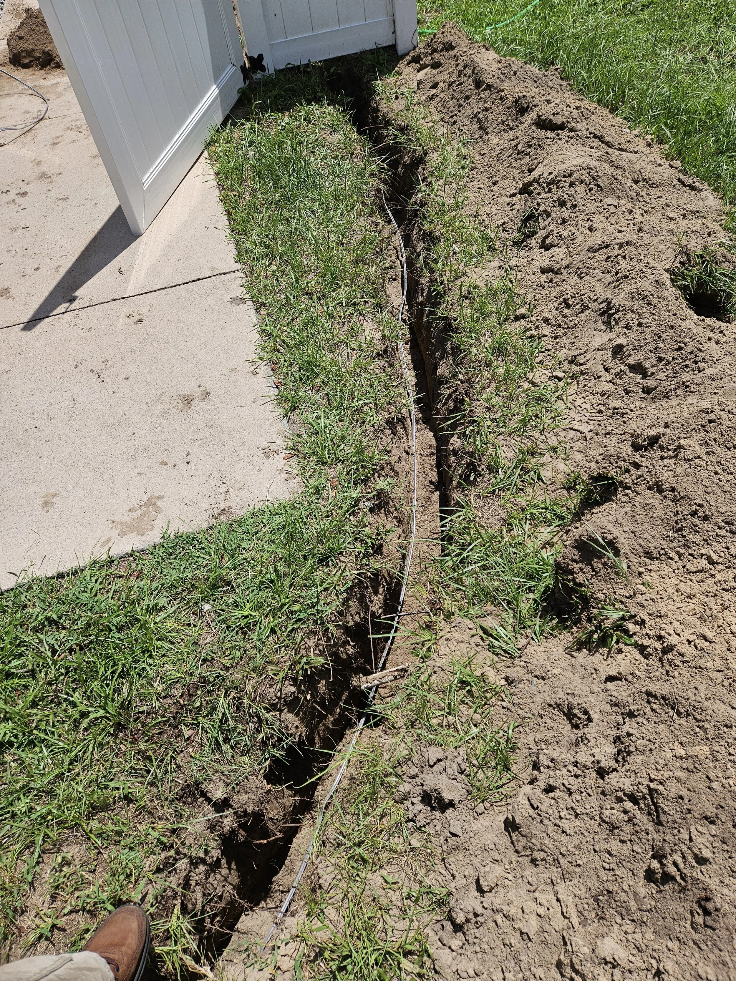 A trench dug in the ground next to a house, with a wire line running through the trench, and grass and soil on either side.