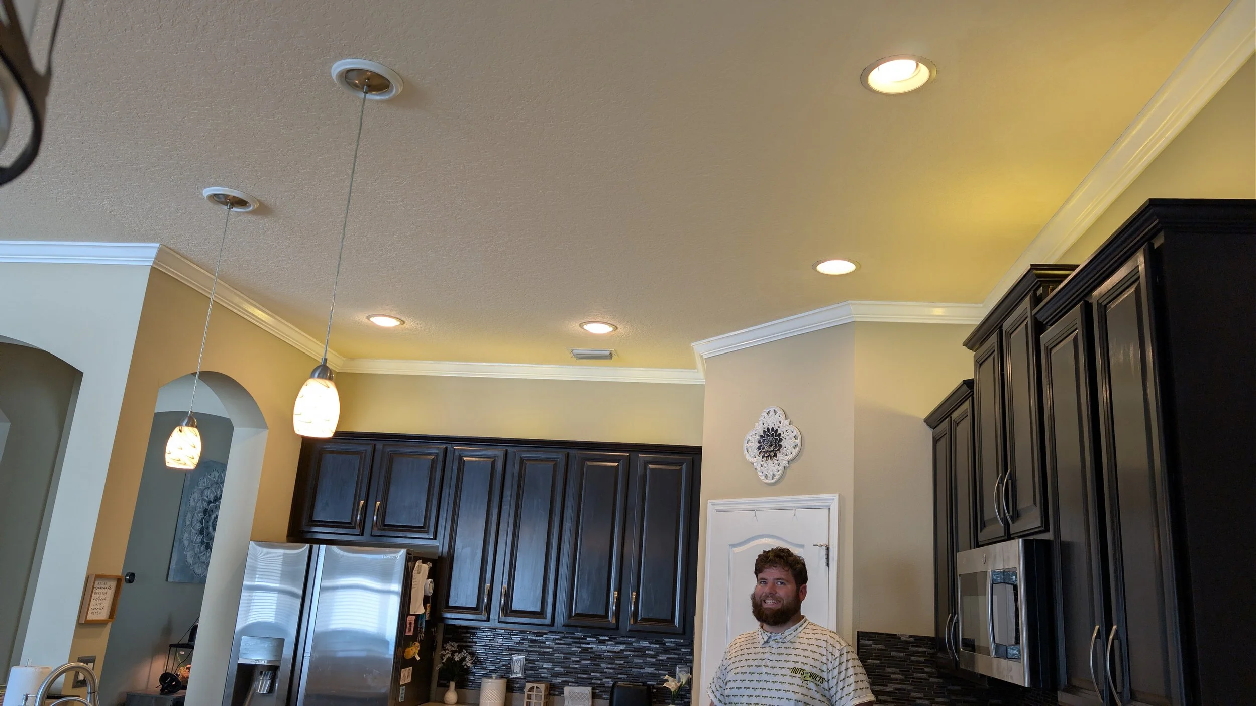 A man with a beard smiling in a kitchen with dark wood cabinets, a stainless steel refrigerator, a microwave, and hanging pendant lights.