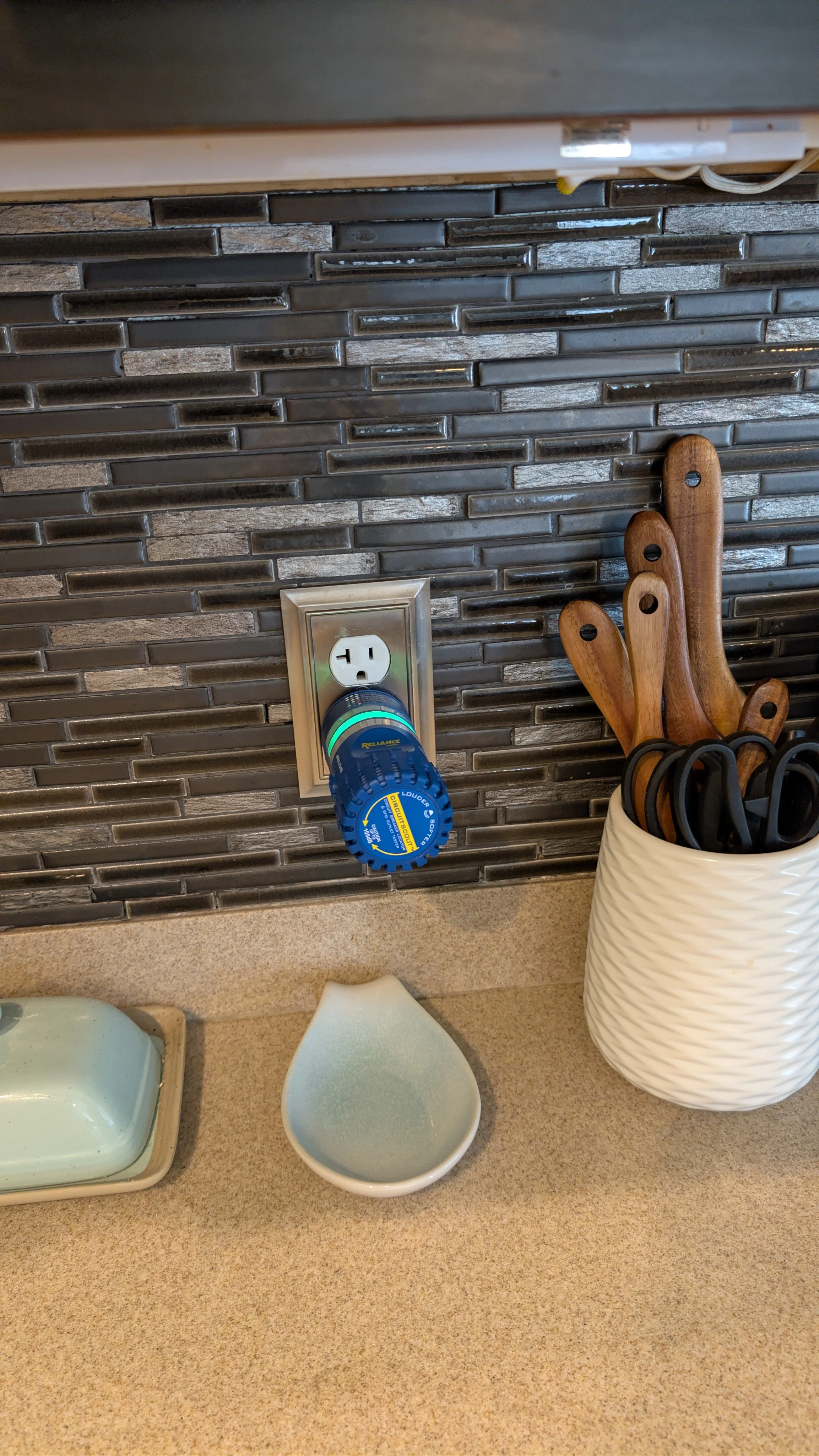 Plug with a blue lock box attached, inserted into an electric outlet on a kitchen backsplash, next to kitchen utensils in a white container.