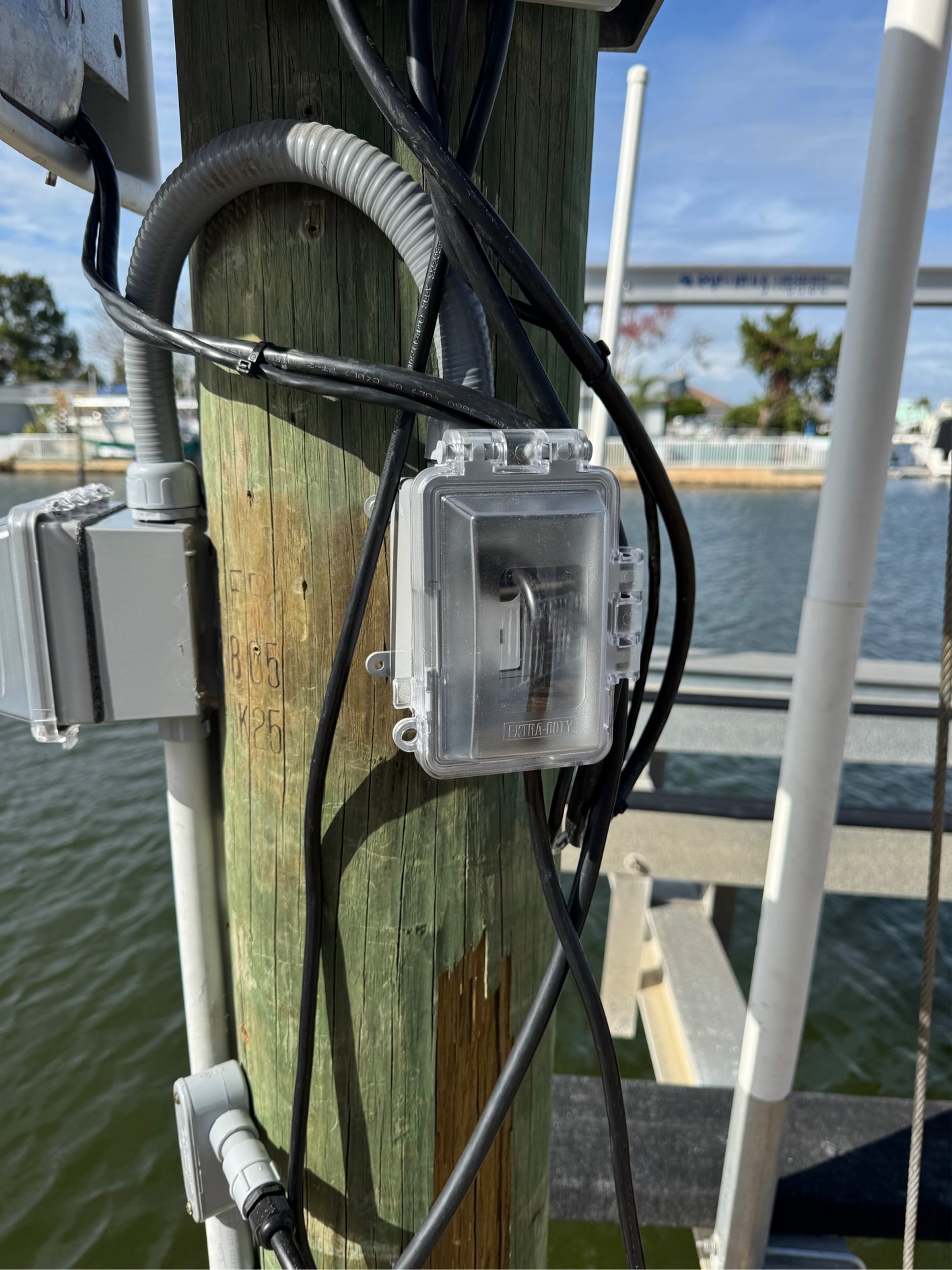 Electrical wiring and boxes attached to a wooden dock post near a waterway with boats and trees in the background.
