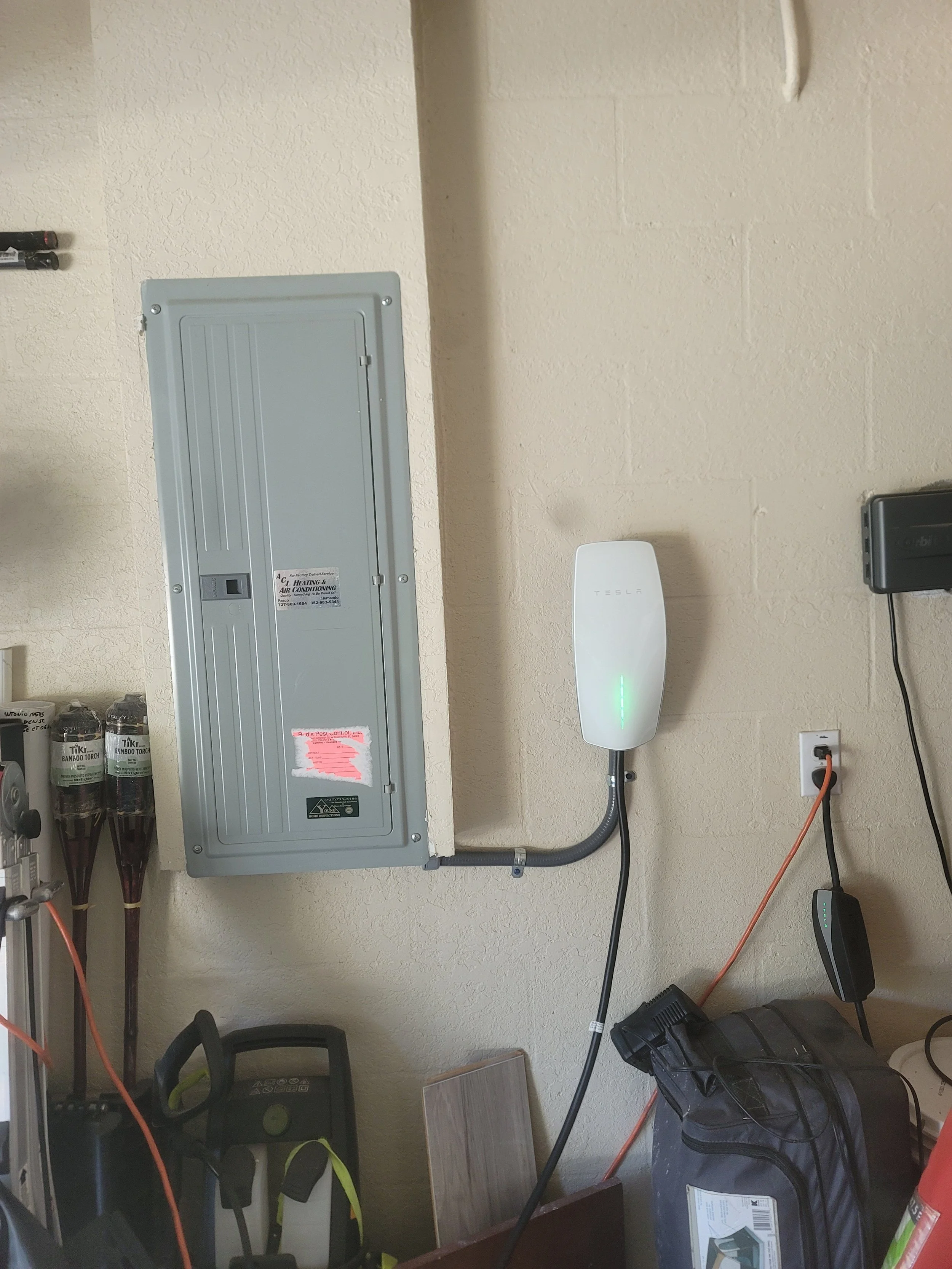 Electric vehicle charging station in a garage, with a Tesla charger plugged into an electric car and other tools and equipment nearby.