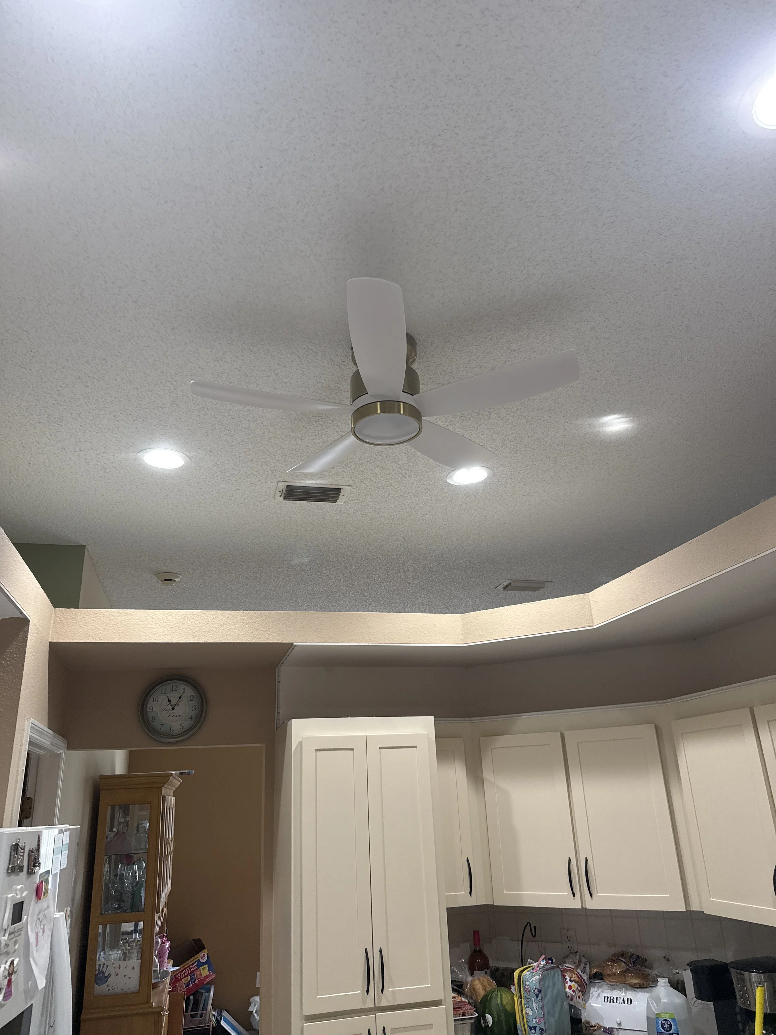 Ceiling fan with three blades in a kitchen with white cabinets and a clock on the wall.