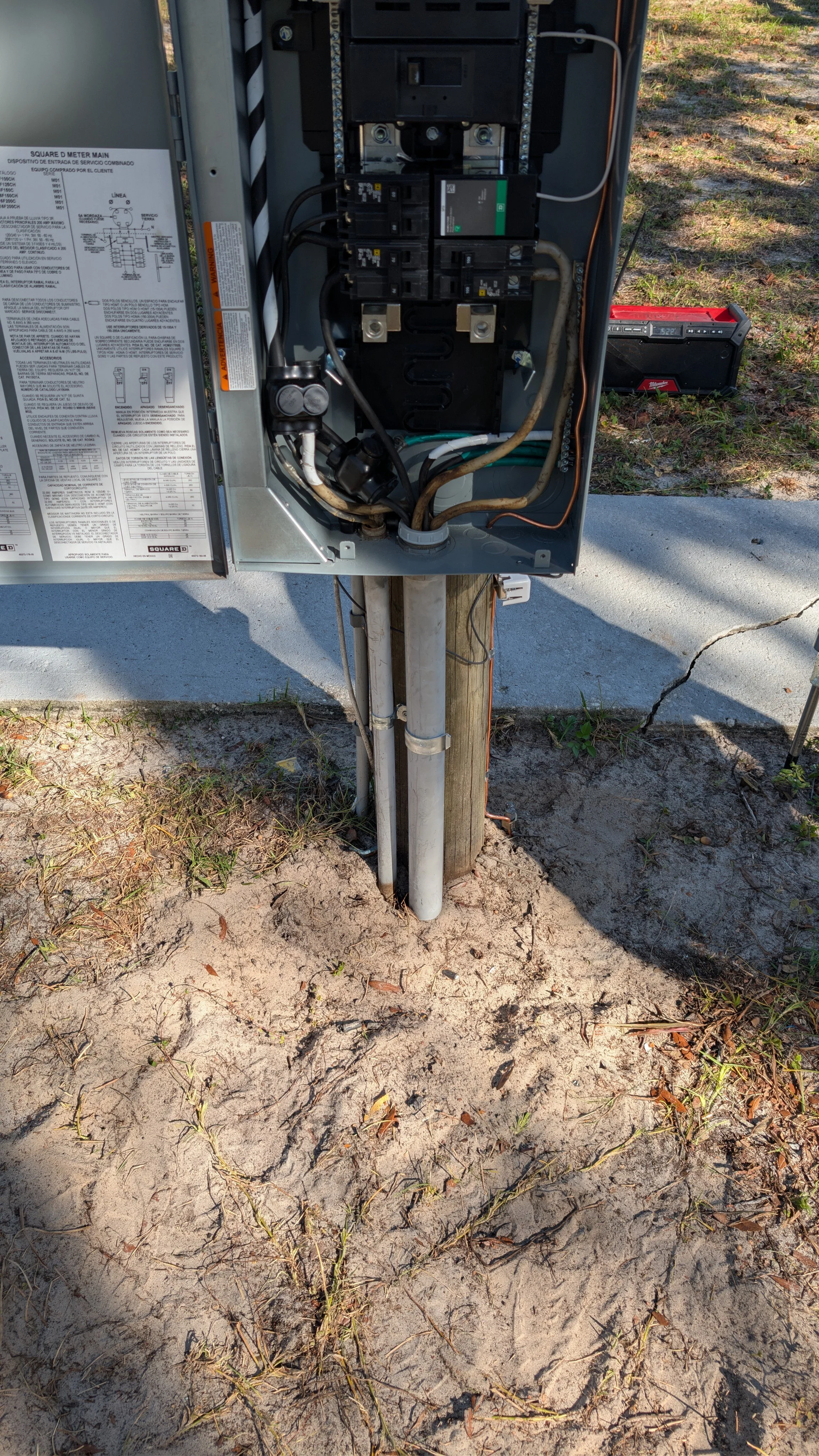 Open electrical panel on a utility pole with visible circuit breakers and wiring, outside on sand and dirt ground.