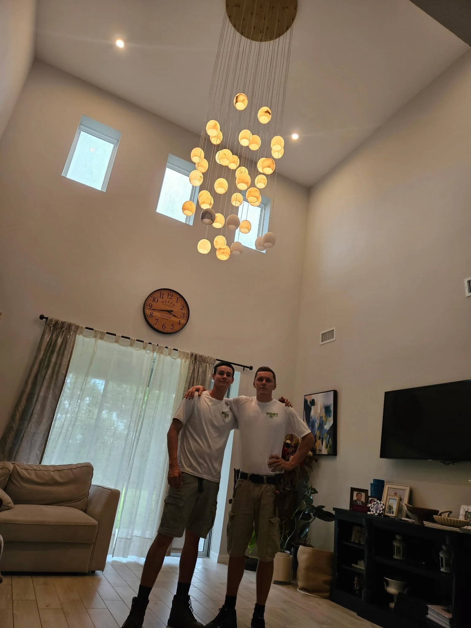 Two young men standing inside a living room with high ceiling, chandelier, and three small windows, posing with arms around each other's shoulders.